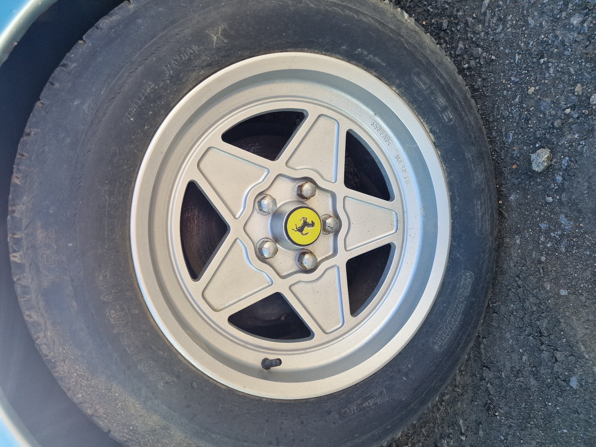 The image presents a close-up view of a car's wheel, featuring a tire mounted on a five-spoke alloy rim. The vehicle is stationary on a surface composed of asphalt and small gravel, under clear daylight conditions. A sliver of a blue car body is visible in the upper left corner.
The tire is black and exhibits signs of age and wear, including fine cracks on the sidewall. Prominently visible text on the tire sidewall includes "RADIAL X" and "TRX," identifying it as a Michelin TRX radial tire. The size specification "240/55VR 415" is clearly readable, denoting a 240mm section width, 55% aspect ratio, V speed rating, and a unique 415mm rim diameter (approximately 16.3 inches). Other partial text includes "TUBEL***S" (likely "TUBELESS"), "MAX LOAD", and "MADE IN FRANCE".
The rim is a light silver or grey color, designed with five spokes forming a star pattern. At its center, a yellow cap displays the iconic black prancing horse logo of Ferrari. The word "Ferrari" is embossed or engraved on one of the spokes, and the rim size "160 TR 415" is also visible, confirming the 415mm diameter matching the tire. Several lug nuts secure the wheel, and a black valve stem is present. The distinct tire and rim specifications, coupled with the Ferrari branding, suggest this is a wheel from a classic Ferrari model, commonly found on vehicles from the late 1970s and 1980s. No individuals or specific activities are depicted. The scene is an outdoor setting, consistent with its provided location context of South Tyneside, United Kingdom.