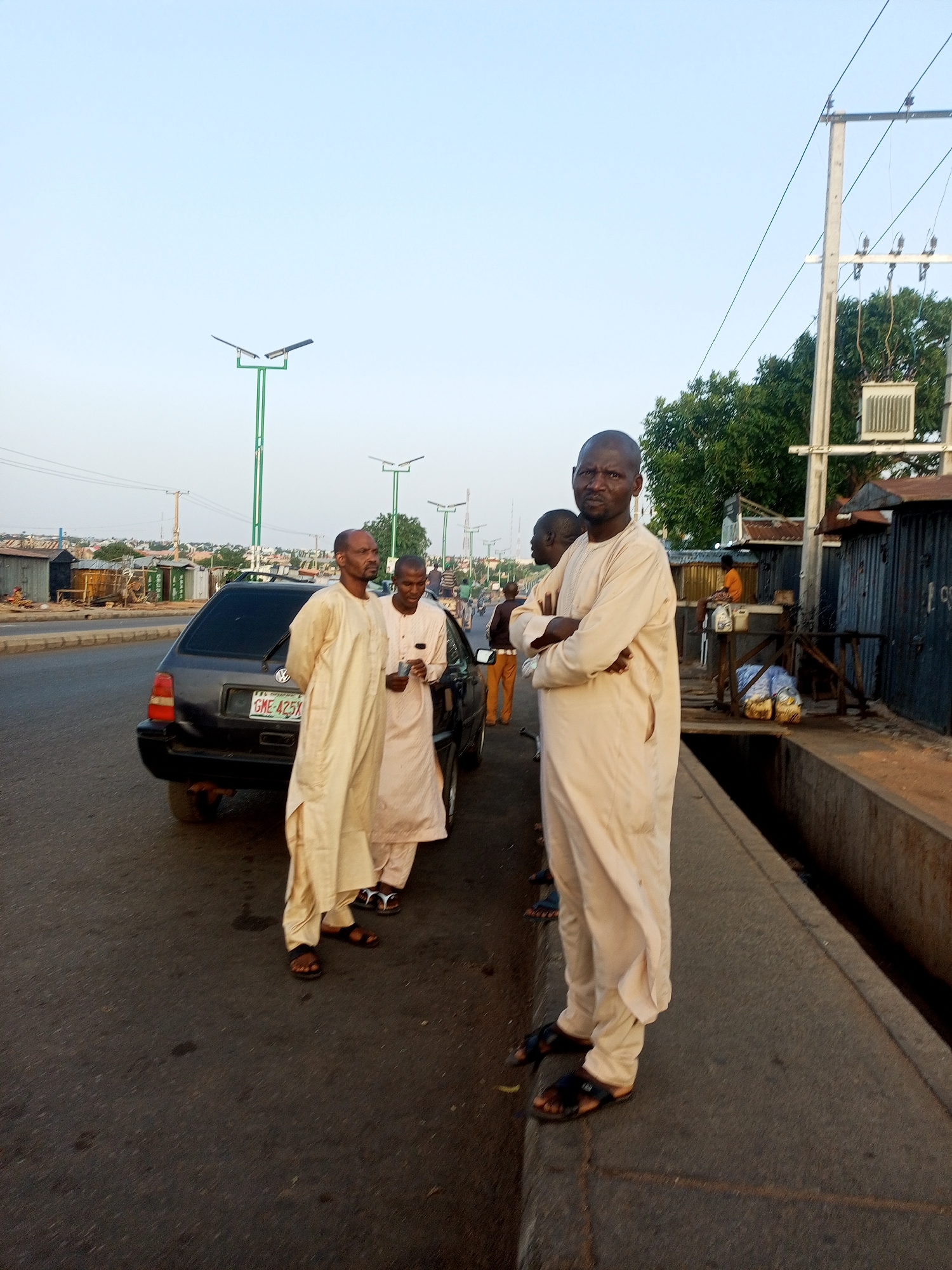 The image shows a street scene with several individuals. In the foreground, a man wearing a light-colored robe and sandals stands with his arms crossed, facing the camera. Behind him, two other men in similar robes stand near a dark-colored car with the license plate "GME 425X." Another person in orange pants is seen in the background, possibly walking away. The setting appears to be urban, with overhead power lines, street lights, and commercial structures lining the road. The location is the city of Unknown, Unknown.