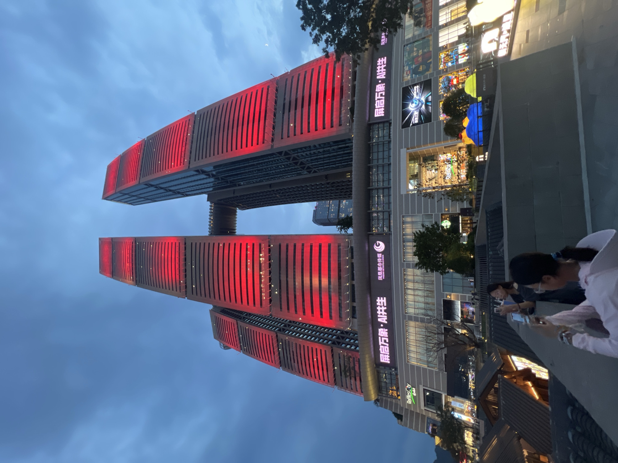 This image captures the Raffles City Chongqing complex, located in the Yuzhong District of Chongqing, China. The iconic twin skyscrapers, illuminated with vibrant red lighting on their upper sections, dominate the skyline. A connecting skybridge spans between the two towers. Below the towers, a multi-story commercial building displays various advertisements and storefronts, including "Raffles City" signage. In the foreground, two individuals are visible. One person, wearing glasses and a dark top, stands on a raised platform, holding a beverage and looking towards the right. Another person, in the foreground and partially obscured, is wearing a pink top and appears to be holding a phone, possibly taking a photo. The scene is set against a twilight sky with visible clouds, and the lower levels of the buildings show a more traditional architectural style with tiled roofs. The overall impression is a modern urban landscape at dusk, highlighting the architectural marvel of Raffles City.