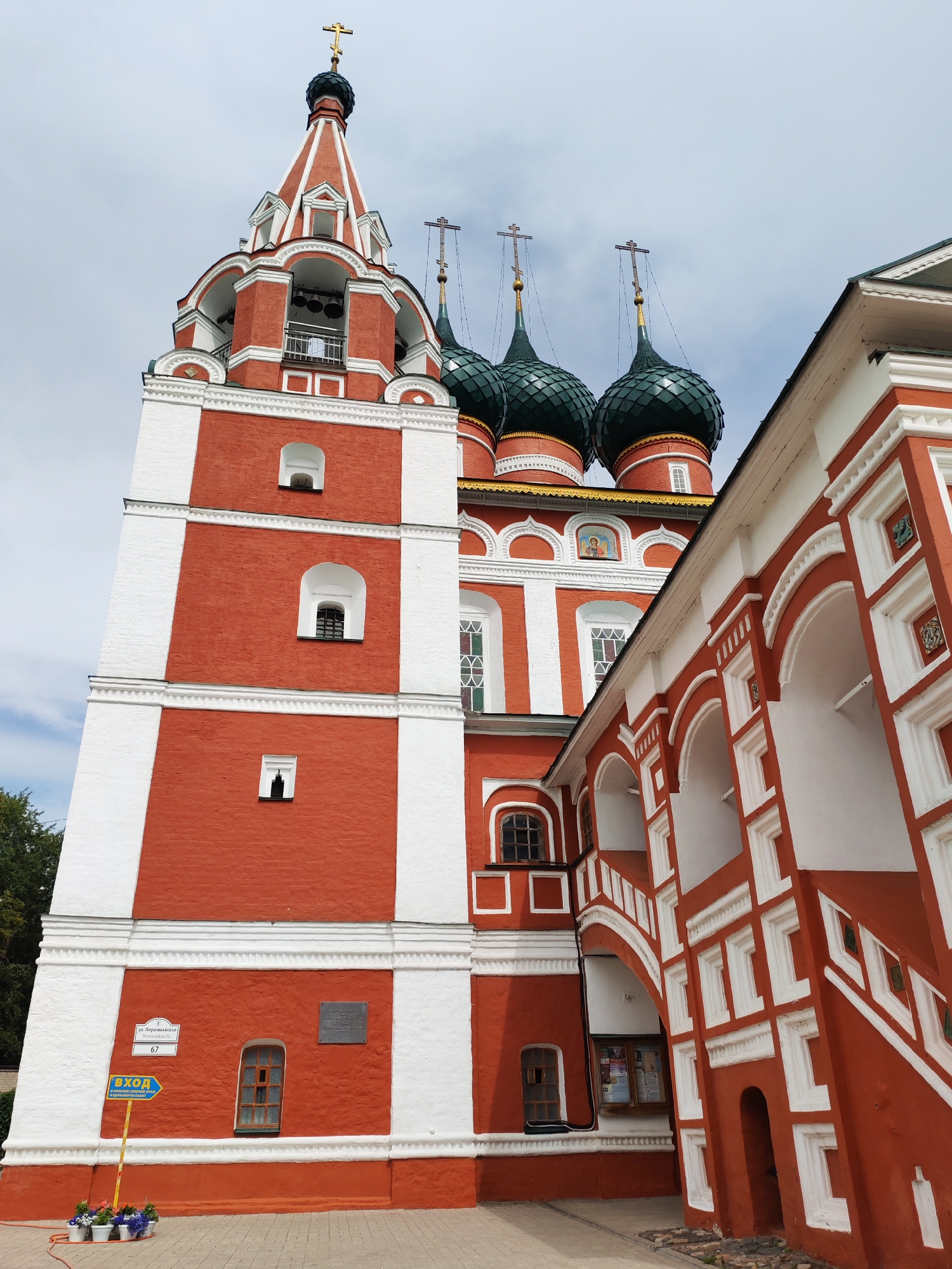This is a low-angle outdoor shot of a brightly colored Orthodox church with a bell tower. The church has red brick walls with white trim. The bell tower is tall and slender, with multiple levels and arched openings. It is topped with a red and white spire and a golden cross.
Adjacent to the bell tower are three green onion domes, also topped with crosses. The building to the right of the bell tower features a white arched walkway with intricate red and white decorative elements. Some windows have stained glass. The sky is overcast, suggesting a cloudy day.
Near the base of the bell tower, a blue directional sign points to the left and reads "ВХОД" (Entrance) in yellow text. Below this, smaller text is present but illegible. Another sign on the wall above the entrance reads "ул. Первомайская" (Pervomayskaya Street) and "д. 67" (building 67). A dark plaque with writing is also visible on the red wall. A few potted plants are at the base of the sign. The scene appears to be a calm, possibly tourist-oriented, setting in Rusanovo, Russia. No people are visible in the frame.
