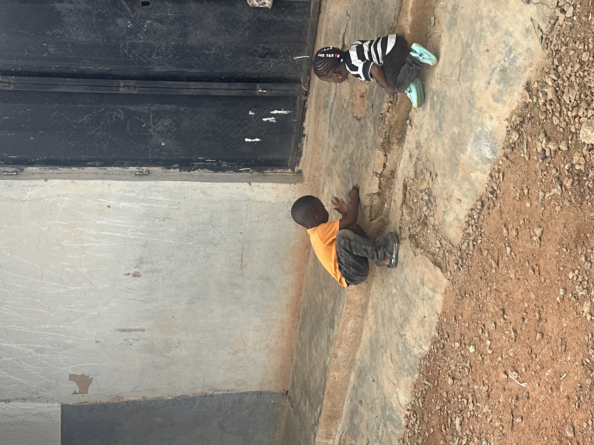 In Gura Top, Nigeria, two young children are crouched on a concrete ledge in front of a weathered building. The child on the left, wearing an orange t-shirt and dark pants, is looking down and appears to be interacting with something on the ground. The child on the right, dressed in a black and white striped shirt and dark pants, is also looking down, with light blue footwear on. Behind them, a large, dark metal door is visible, and the wall of the building shows signs of wear and tear, with peeling paint and scuff marks. The ground in front of the ledge is covered with small rocks and reddish-brown soil.