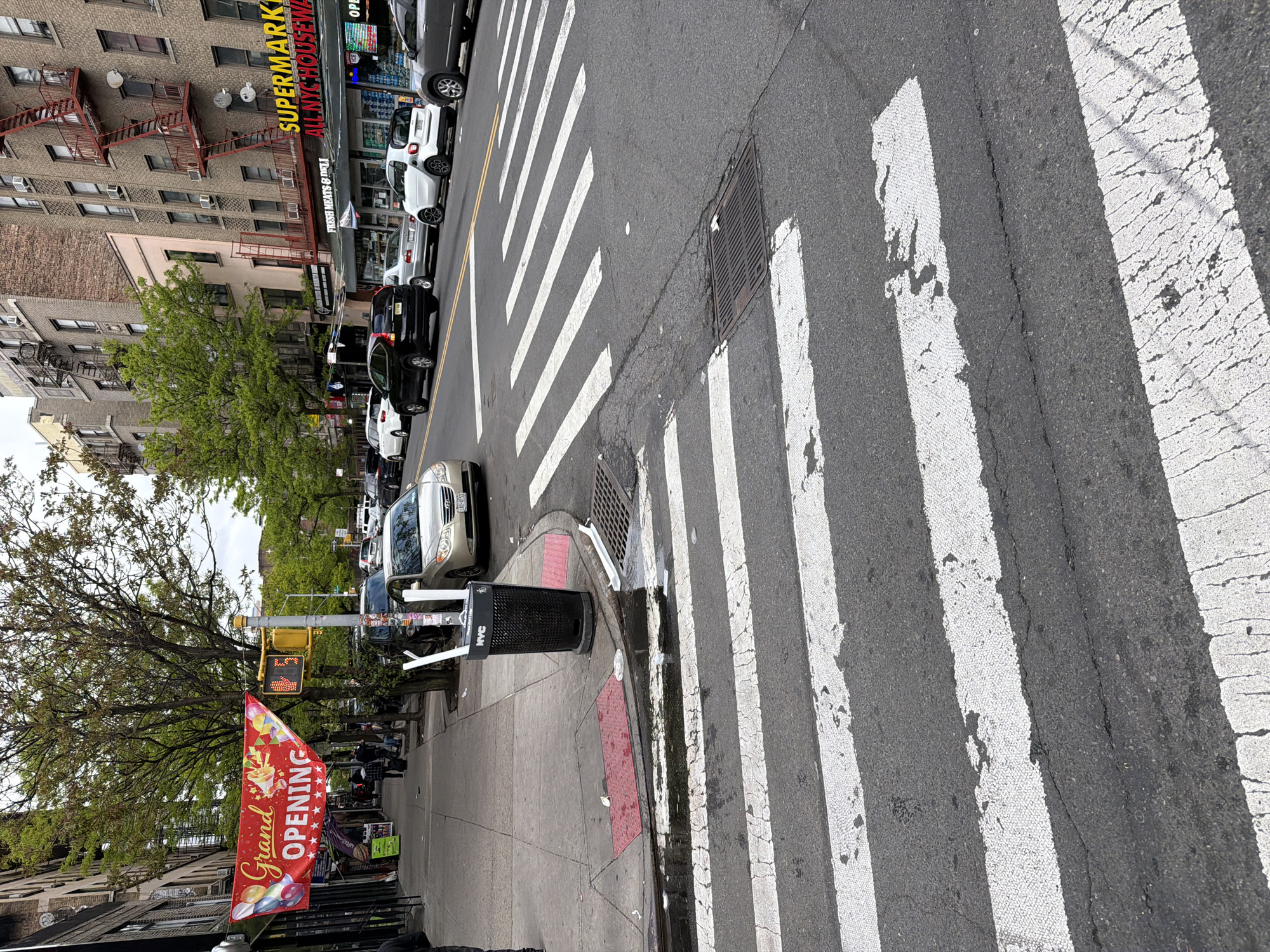 This image, rotated 90 degrees clockwise, presents a daytime street scene in what is identifiable as New York City. The perspective is from the sidewalk, looking across a busy street toward buildings and parked cars.
In the foreground, a prominent white pedestrian crosswalk stretches across a grey asphalt road, displaying visible cracks and wear. A metal storm drain grate is embedded in the street near the curb. On the concrete sidewalk to the left, a black public trash can with the "NYC" logo stands next to a red "Grand Opening" banner, decorated with balloons and confetti, indicating a new business. Above the banner, a yellow traffic light pole holds a pedestrian signal showing a countdown of "25".
The midground features a line of cars parked along the curb, with various models and colors visible. Behind the sidewalk and cars, several trees with full green foliage suggest a spring or summer setting. A few pedestrians can be seen walking on the sidewalk further down the block.
The background is filled with multi-story urban buildings, typical of New York. One building is made of reddish-brown brick and features external metal fire escapes. Below it, a storefront sign clearly reads "SUPERMARKET" and "ALL NYC HOUSEWARE" in bright yellow letters. A smaller sign beneath it partially shows "Fresh MEATS & DELI". The lighting appears diffuse, suggesting an overcast day or soft daylight. The scene depicts a lively urban environment with a mix of residential and commercial activity, and the infrastructure shows signs of age and constant use.