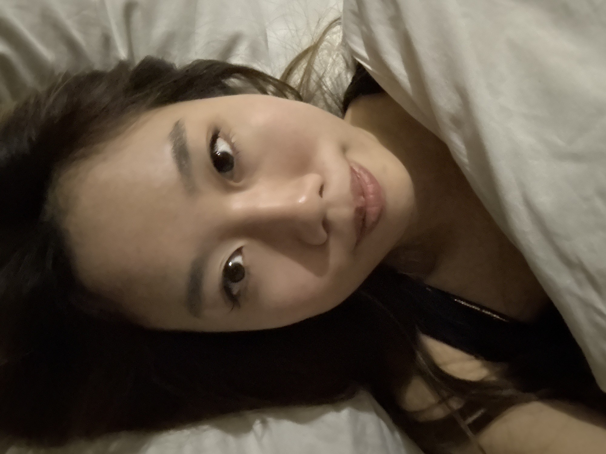 The image is a close-up selfie of an Asian woman lying in bed. She is partially covered by a white blanket. Her hair is dark brown and shoulder-length. She has a slight smile. The background appears to be a bedroom setting. There is no information to support this image being taken in Dunhuang City, China.