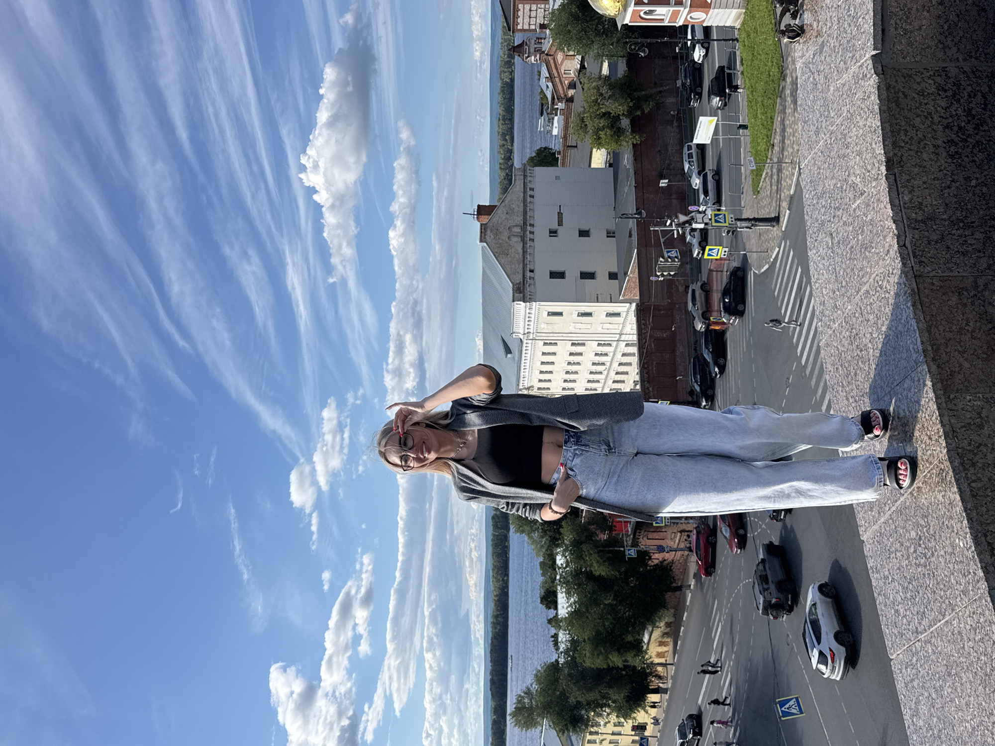 The image shows a young woman standing on a stone ledge, looking at the camera and making a peace sign with her hand. She is wearing a grey cardigan over a black crop top and light blue jeans. She has blonde hair, glasses, and is wearing sandals.
In the background, there is a body of water, a cityscape with buildings and trees, and a street with cars and pedestrians. The sky is blue with white clouds, suggesting it is daytime and the weather is fair. The overall mood of the image is casual and cheerful. The scene appears to be an urban setting with a waterfront.
There is some visible text on a blue diamond-shaped sign on the street which appears to be a traffic sign. The location context is Novokuybyshevsk, Russia.