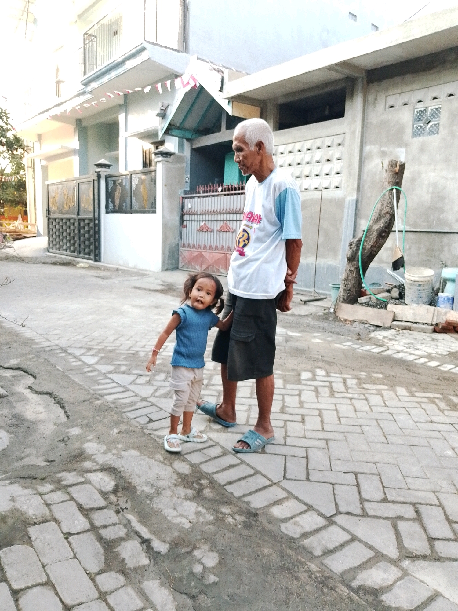 In this medium shot, a young girl and an elderly man stand on a paved walkway in front of a building. The girl, with dark hair and wearing a blue t-shirt and light-colored pants, stands to the left, looking towards the camera. Her right hand is by her side, and her left hand is near the man's leg. She wears white sandals. The man, with white hair and a light blue and white t-shirt, stands to the right. He wears dark shorts and blue sandals. His left hand is behind his back, and his right hand is holding the girl's arm.
The background shows a building with a grey concrete wall and a tiled roof. Small Indonesian flags are strung along the overhang of the building. The walkway is paved with rectangular bricks. To the right of the man, a tree trunk stands with a green hose wrapped around it, and some buckets and construction materials are visible at its base. The overall scene suggests a casual outdoor setting, likely in the daytime due to the bright lighting. The emotions appear neutral, with the girl looking curious.