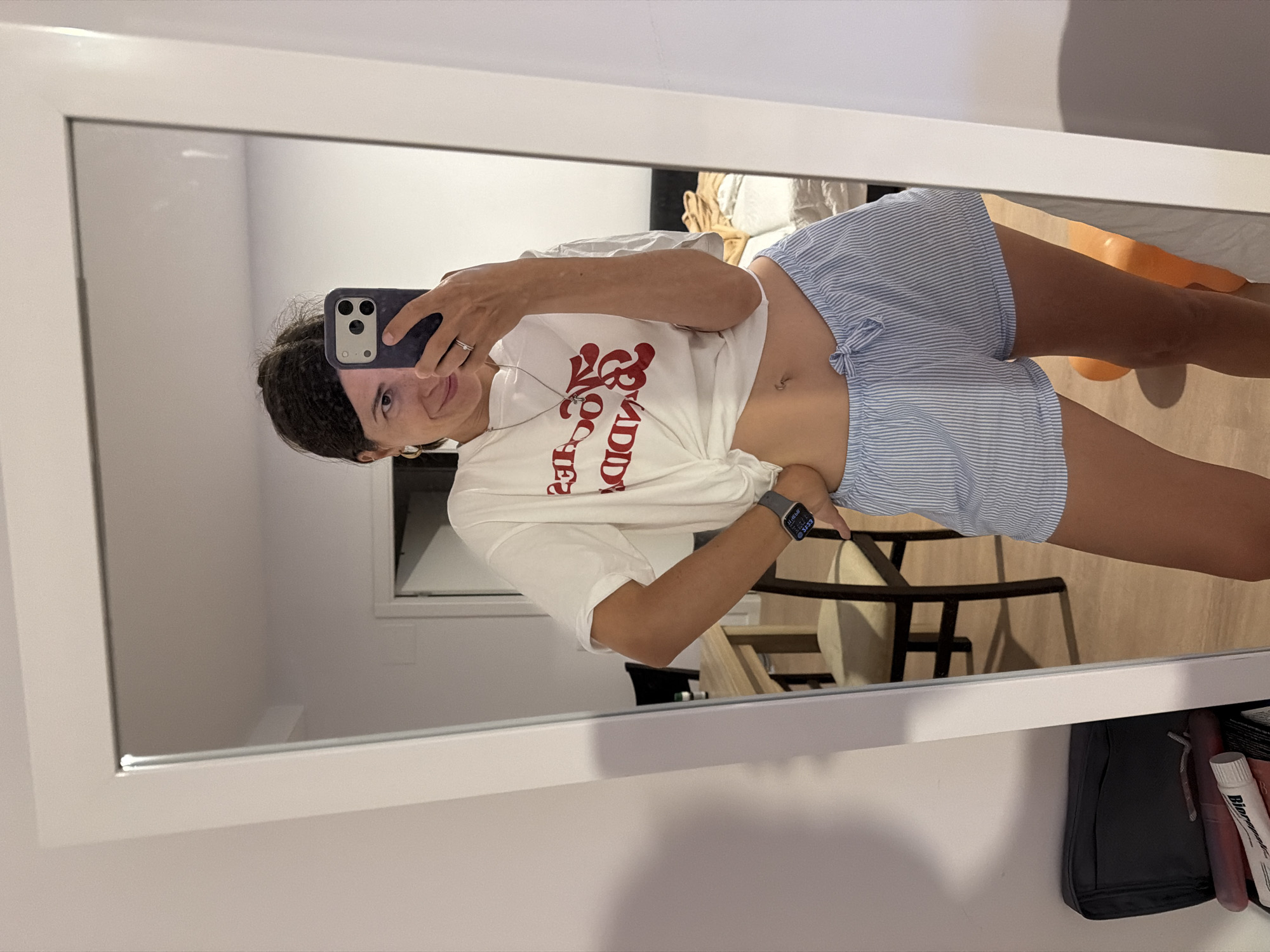 A woman is taking a mirror selfie in what appears to be a bedroom. She is wearing a white t-shirt tied at the waist, a necklace, and blue and white striped pajama shorts. She has a watch on her left wrist and a ring on her right hand. In the background, a bed and a chair are visible. The overall setting suggests a domestic environment, possibly an apartment in l'Hospitalet de Llobregat, Spain, given the provided context.