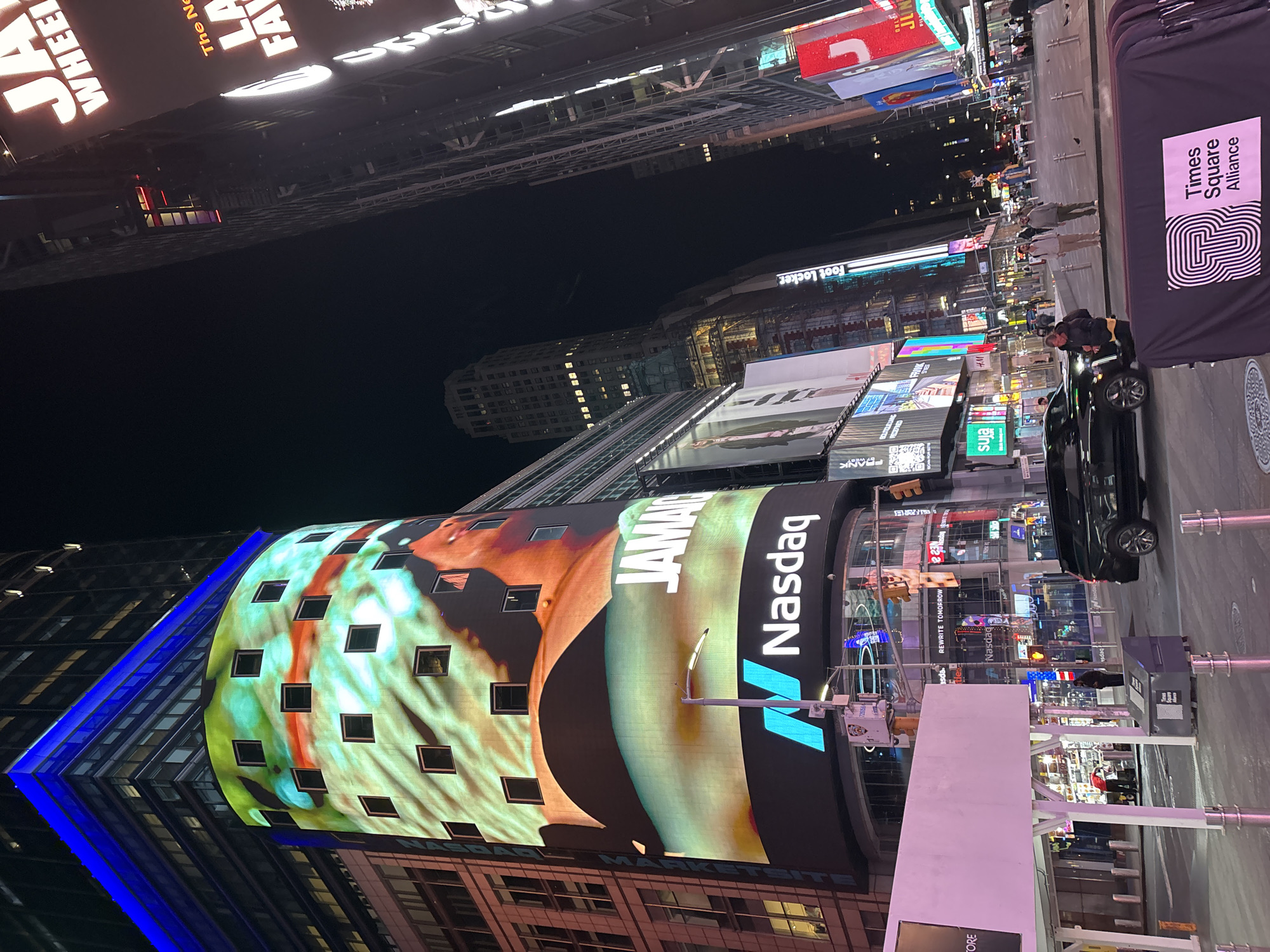 This night-time image captures the iconic, brightly lit urban landscape of Times Square in New York City, USA. The scene is dominated by towering buildings adorned with numerous vibrant digital billboards.
The most prominent feature is the curved facade of the Nasdaq MarketSite building on the left, showcasing a massive digital screen. This screen displays a colorful advertisement featuring an abstract image of a person, the partially visible word "JAMA" (likely part of "JAMAICA"), and the large "Nasdaq" logo. Below this, another sign reads "REWRITE TOMORROW".
The street level in the foreground shows a few pedestrians and a dark-colored car. To the right, a black banner with the "Times Square Alliance" logo and text is visible. Various other illuminated advertisements cover the surrounding buildings, displaying brand names such as "Foot Locker," "HM" (H&M), and "suja." Partially visible text from higher billboards includes "J WHEEL," "The Ne," and "LA FA," suggesting ads for attractions like "The New York Wheel."
The time of day is clearly night, with the dark sky contrasting sharply with the intense, artificial illumination from the digital displays. The atmosphere is bustling and energetic, characteristic of Times Square, with constant visual stimulation from the electronic signage.
