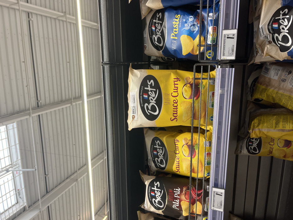 The image shows four bags of Bret's potato chips, on a shelf in a supermarket, located in Saint-Brice-Courcelles, France. Each bag features a different flavor, displayed on the front with a picture of the chips. The first bag features the flavor "Pili Pili", the second bag is "Sauce Curry", the third bag is also "Sauce Curry" and the last bag is "Pastis Sans Alcool". There is a price tag attached to the last bag, indicating a price of 1.70 euros. The background of the image is a white ceiling, with a skylight that appears to be made of glass or plastic. There are no people in the scene.
