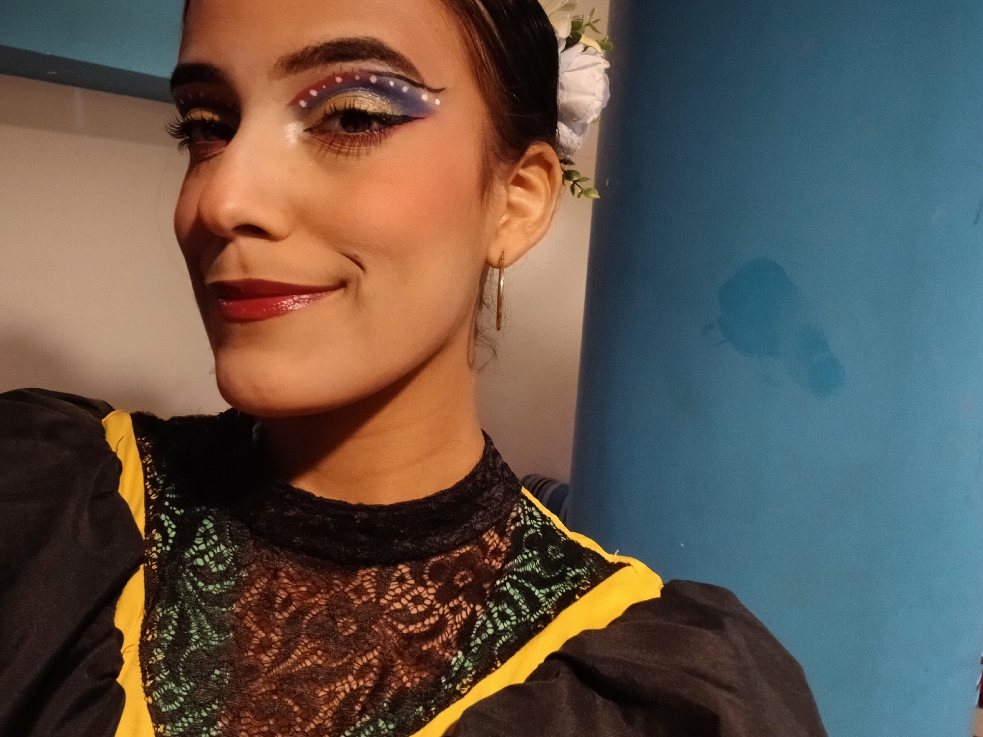 A young woman with elaborate eye makeup is centered in the image. The makeup features a gold base, topped with a blue and red arc outlined with white dots. She has dark hair pulled back, adorned with white flowers. She has hoop earrings. She is wearing a black top with a lace overlay and yellow detailing on the shoulders. The background is a blue wall. The specific location within Playa El Agua, Venezuela, cannot be determined from this image.