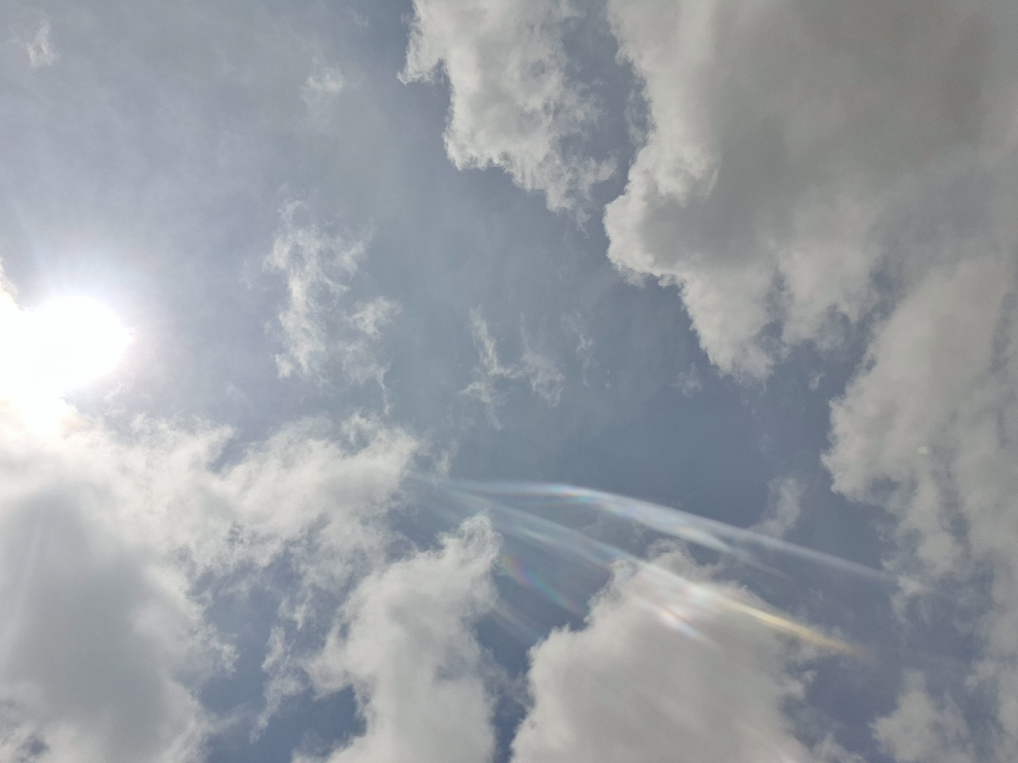 A bright sun is partially obscured by clouds, casting a soft glow and creating visible light shafts that refract into subtle rainbow hues against a pale blue sky. Several fluffy white clouds are scattered across the frame, with larger, more dense formations dominating the lower portion of the image. The overall impression is of a bright, slightly hazy day. The location is Gura Top, Nigeria.