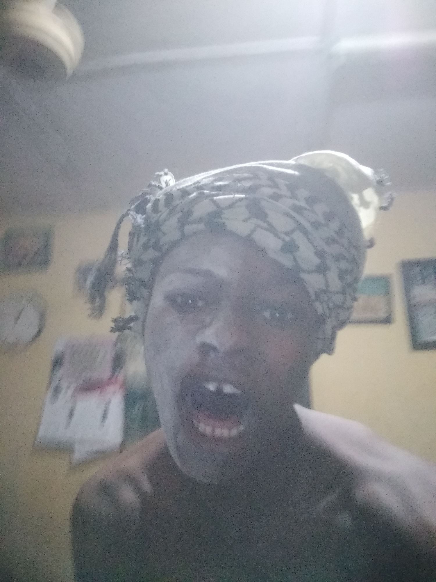 A man wearing a head covering is pictured in an indoor setting. The location appears to be a room with light-colored walls, decorated with a calendar and framed pictures. The man is close to the camera, with his mouth open. A ceiling fan is partially visible in the upper left corner.