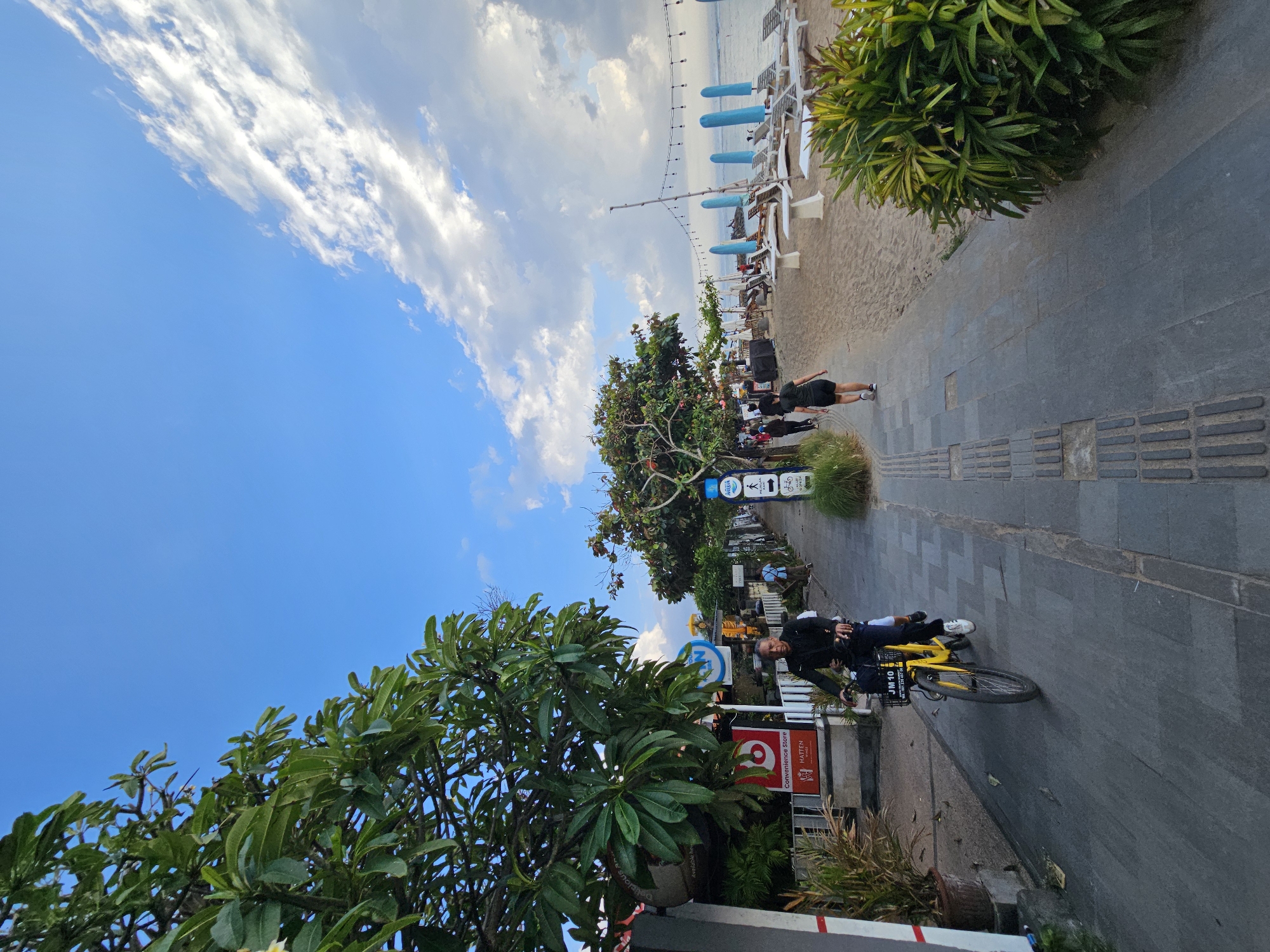 This image depicts a bustling pedestrian walkway adjacent to a sandy beach in Denpasar, Indonesia. On the left, a man rides a yellow bicycle with a basket containing what appears to be a young child or person in the back. He is wearing a dark long-sleeved shirt and dark pants, with white sneakers. The walkway is paved with dark gray stones and features tactile paving for the visually impaired. Several trees with lush green foliage line the left side of the walkway, along with planters and commercial signage for a convenience store.
To the right of the walkway is a wide expanse of sand with rows of white lounge chairs and large blue beach umbrellas. In the distance, the ocean is visible under a bright blue sky dotted with fluffy white clouds. Several string lights are strung overhead along the beach area. Other individuals are seen walking along the pathway and on the beach, suggesting a popular public space. The overall impression is one of a vibrant coastal scene in a tropical urban environment.