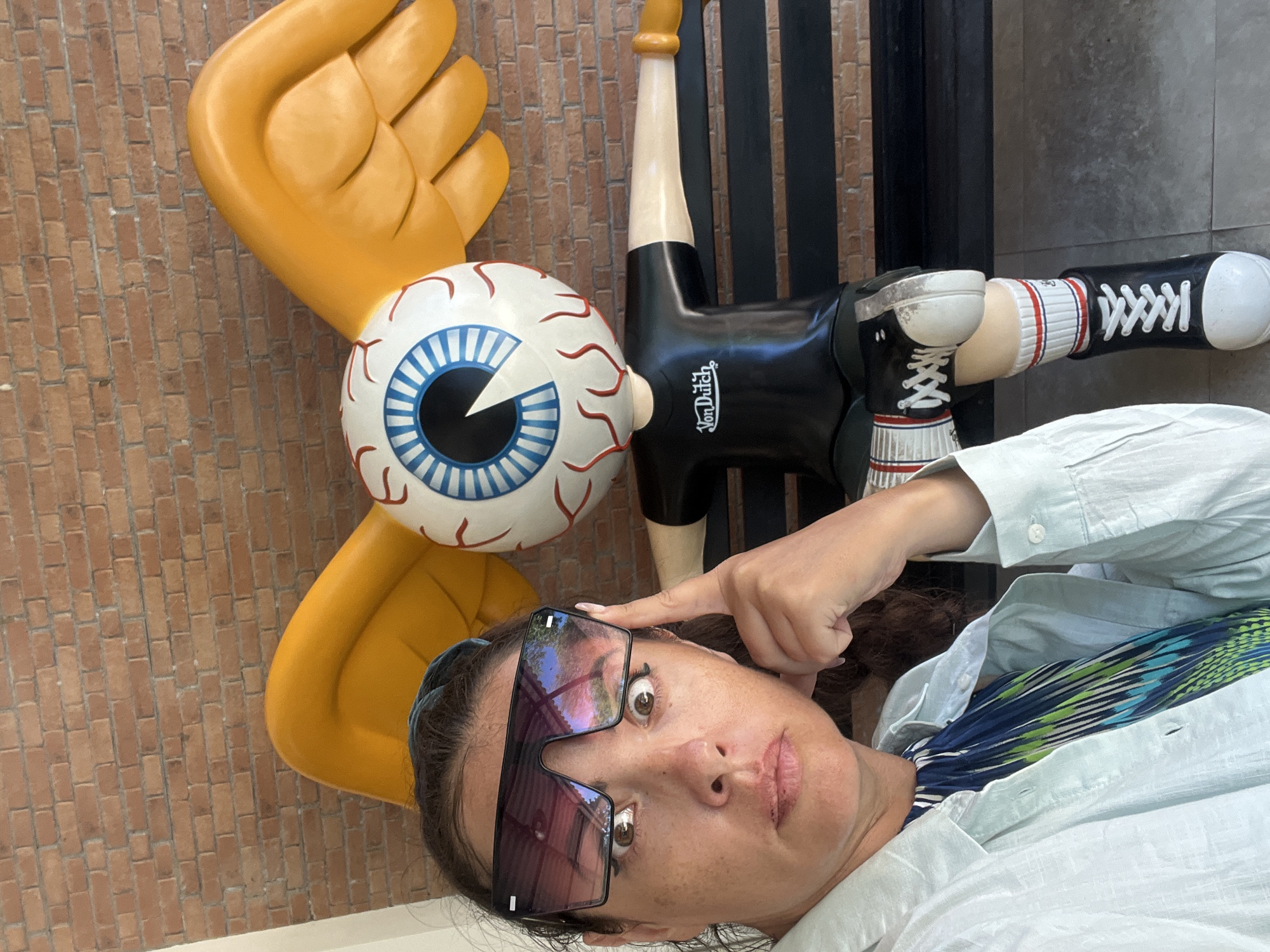 A woman with dark hair and light skin, wearing oversized sunglasses, a light blue button-up shirt, and a colorful patterned dress, is taking a selfie. Her left hand is raised to her temple, and she is looking directly at the camera with a surprised expression. To her right, a large, colorful statue of a figure with an eyeball for a head, yellow wings, and wearing a black t-shirt and sneakers sits on a dark bench. The statue has "Von Dutch" printed on its t-shirt. The background consists of a brick wall. The scene appears to be outdoors. Based on the architectural style and the presence of the Von Dutch brand, which has a presence in Bali, this photo was likely taken in Kuta Selatan, Indonesia.
