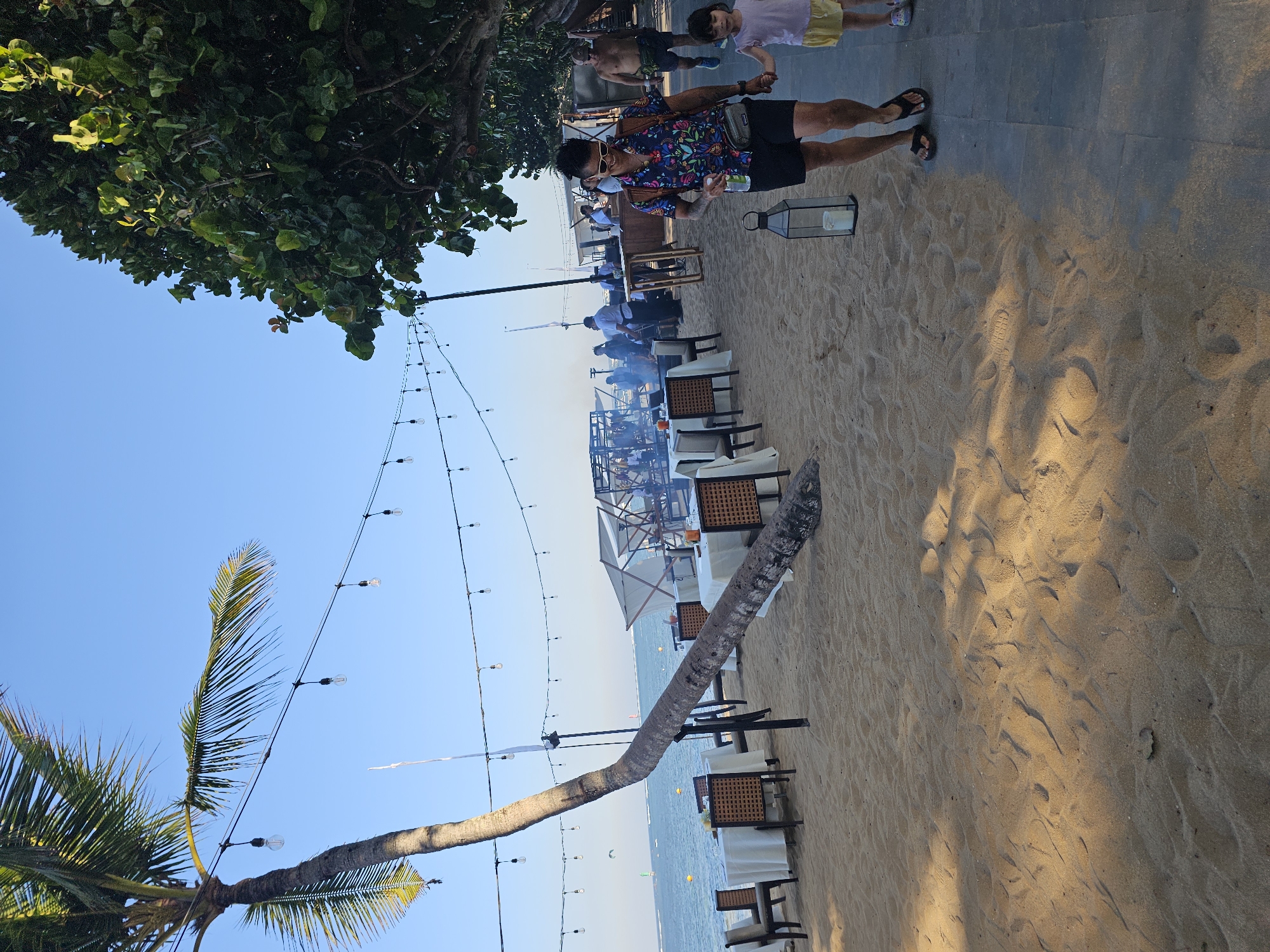 This image depicts an outdoor dining area on a sandy beach in Denpasar, Indonesia, likely within a resort or restaurant. A tilted palm tree with green fronds is on the left, with string lights overhead. In the background, there are tables set with white tablecloths and chairs, and a food preparation area with several staff members visible. To the right, a person wearing a colorful shirt and dark shorts is walking with a young child wearing a yellow dress. The ground transitions from sand to a paved walkway. The ocean is visible in the background, with a clear blue sky above.