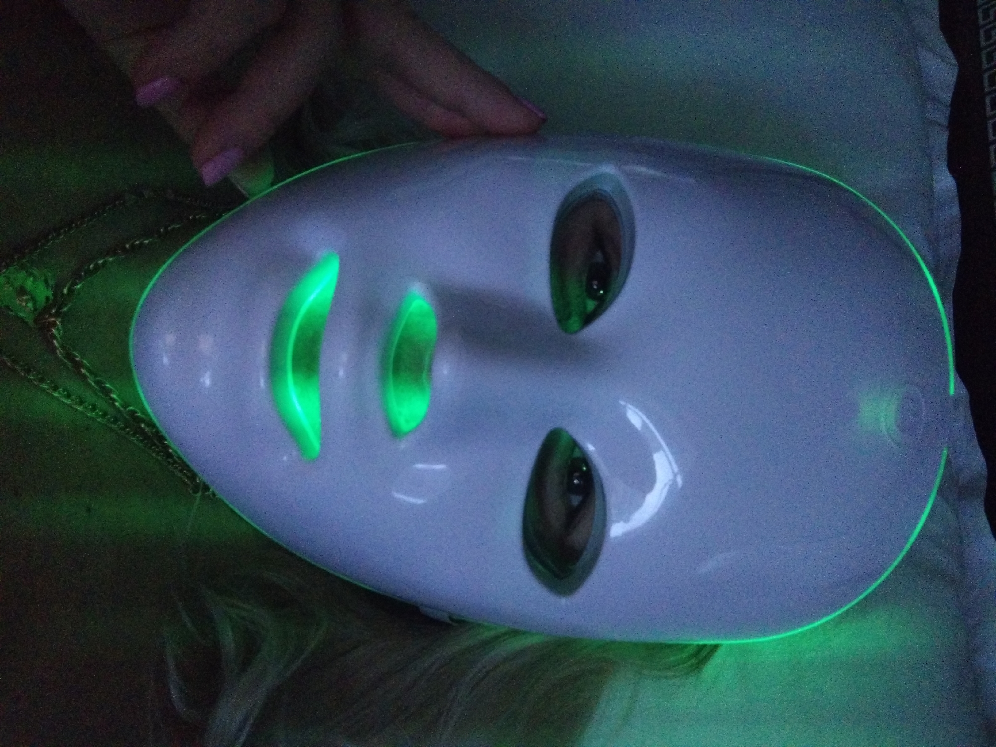 A close-up, eye-level shot shows a person wearing a white LED facial mask with green light emitting from the eye, mouth, and chin areas. The mask covers the entire face, with only the eyes, nose, and mouth visible through cutouts. A hand with a pink manicure is touching the side of the mask near the temple. The person is wearing two layered necklaces. The background is dark and appears to be a bed with some fabric, possibly a pillow, peeking out from the top left corner. The lighting is dominated by the green glow from the mask, casting a diffused green light on the surrounding area. The image does not contain any identifiable landmarks or elements that would allow for pinpointing a specific location within Stara Zagora, Bulgaria.