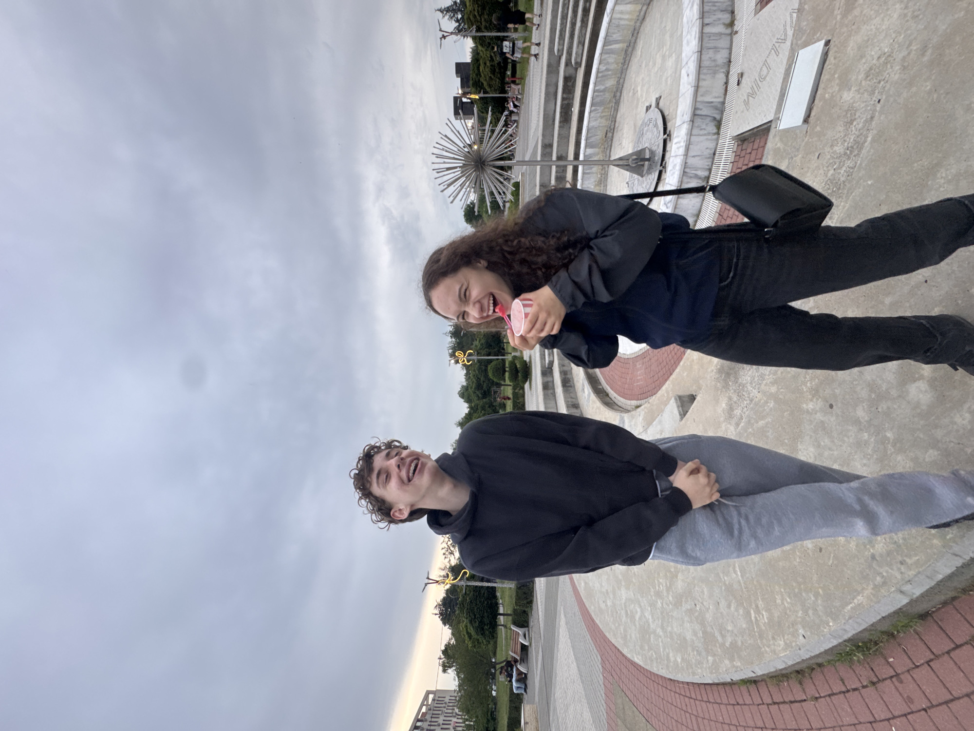 Two young people, a male and a female, are standing outdoors in a park-like setting. The male, on the left, is wearing a black hoodie and grey sweatpants. He is smiling with his head tilted back, revealing braces. His hands are clasped in front of him. The female, on the right, is wearing a dark jacket over a dark top and black jeans. She has curly hair and is holding a pink cup with a spoon, appearing to be eating ice cream or a similar dessert. She is laughing with her mouth open. Behind them, there is a circular fountain with decorative metal spikes, tiered seating, and a walkway with red brick edging. In the background, there are trees, streetlights, benches with people sitting on them, and modern buildings. The sky is overcast with gray clouds.