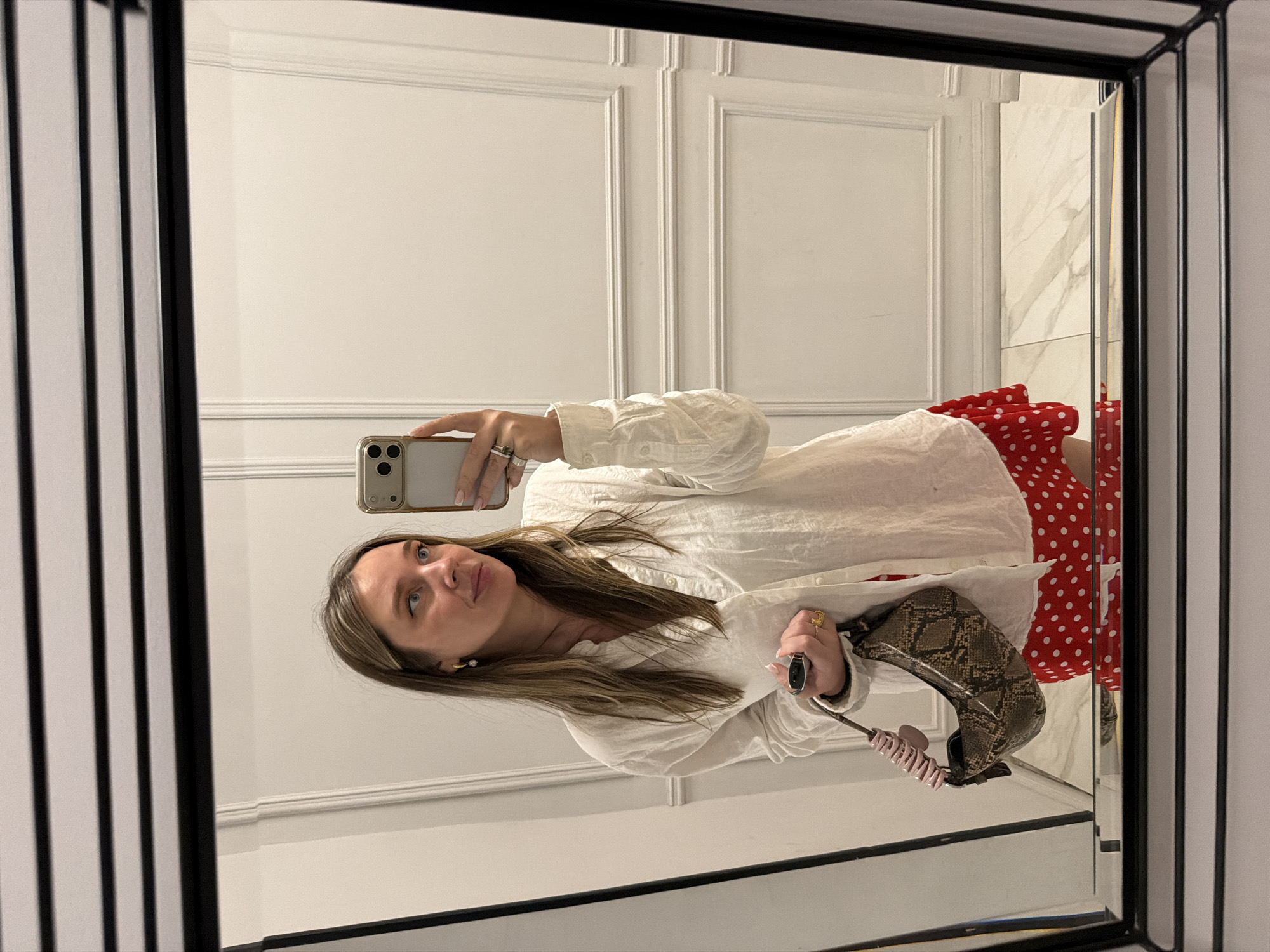 A medium shot depicts a woman reflected in a mirror, taking a selfie. She holds a smartphone in her left hand. The woman has shoulder-length brown hair and is wearing a white blouse and a red polka-dot skirt. In her right hand, she holds a snakeskin-patterned purse and a hair clip. The background features white walls with decorative paneling and a portion of a marble wall visible on the right side. The mirror has a black frame with vertical lines. The location cannot be determined to be Moscow, Russia from the provided image.