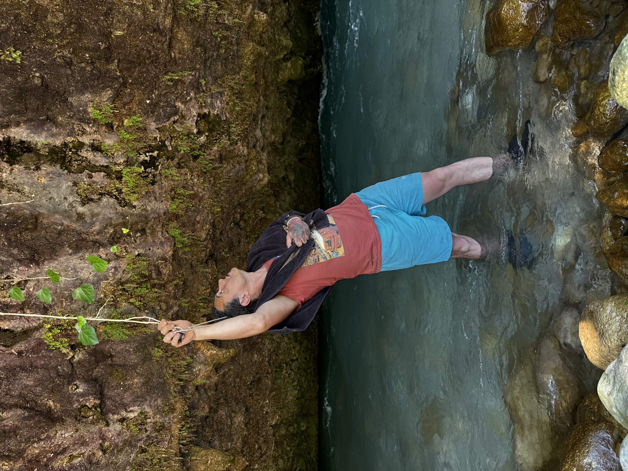 A man stands in shallow, clear turquoise water near a rocky overhang. He is wearing a red t-shirt, blue shorts, and a dark towel draped over his shoulders. His right arm is raised, holding onto a thin rope with several green leaves attached. The water flows gently, with rocks visible on the riverbed and along the bank. The overhang behind him is composed of rough, textured rock with patches of green moss and small plants growing on its surface. The scene appears to be a natural outdoor environment, possibly a river or stream in a forested area. The specific location in Abgara, Abkhazia is not discernible from the image itself.