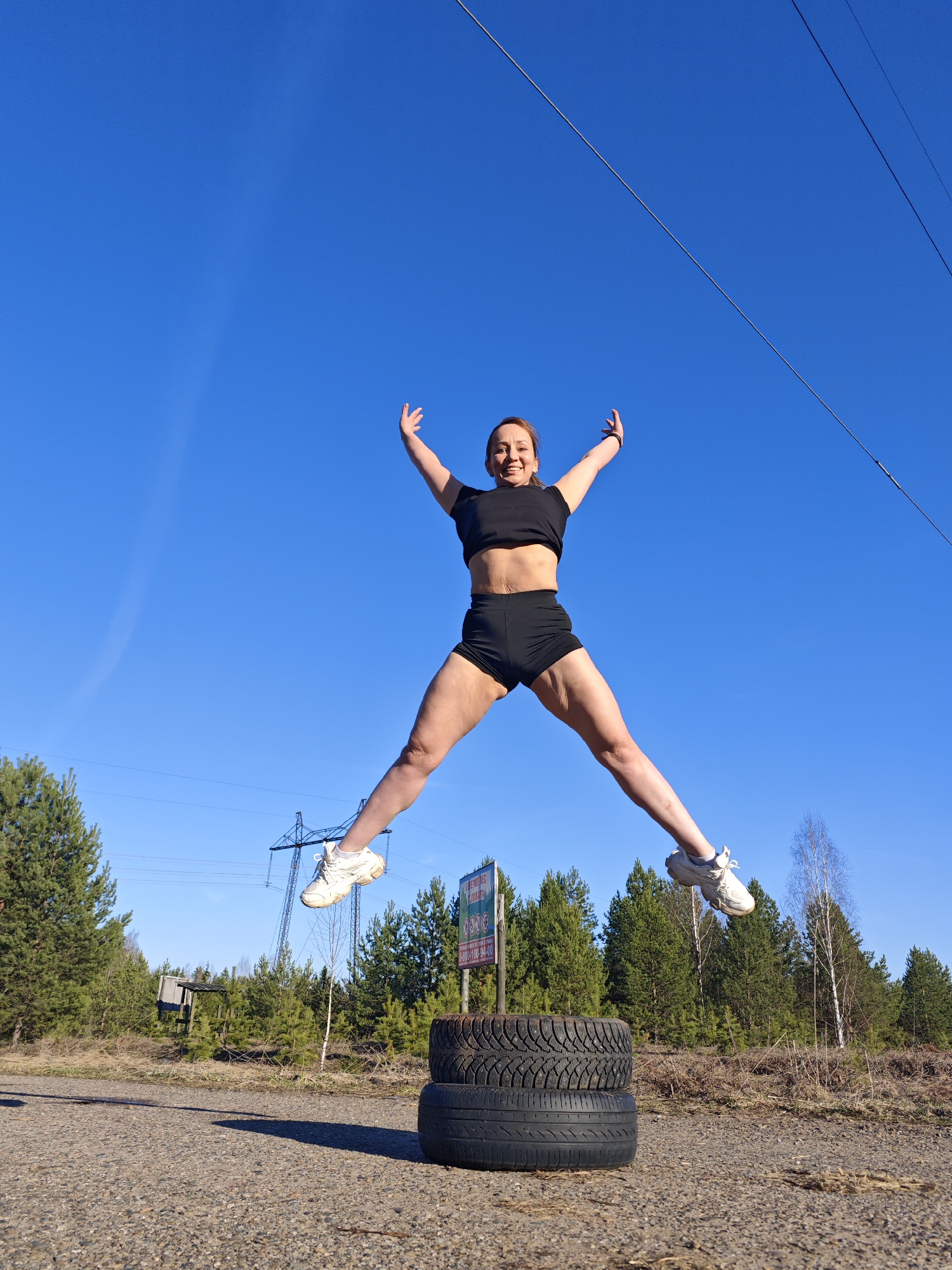 A woman is captured mid-air in a dynamic jump, set against a clear, vibrant blue sky on a sunny day. She appears to be in her 30s or 40s, with a cheerful expression, smiling broadly. She is dressed in black athletic wear, consisting of a short-sleeved crop top and shorts, paired with white athletic shoes. Her arms are raised and spread wide, mirroring her legs which are extended outwards in a star-jump pose, conveying energy and happiness.
Below her, two black tires are stacked on a gravelly ground, positioned almost directly underneath where she is jumping. The setting is an outdoor, possibly rural or roadside area near Chaikovsky, Russia. In the background, a dense forest of coniferous trees, interspersed with some bare deciduous trees, lines the horizon. Power lines and a metal lattice power pylon are visible, stretching across the sky, suggesting an industrial or utility presence in the natural landscape. A distinct shadow of the woman and the tires is cast on the ground, indicating bright sunlight.
To the right, partially visible behind the trees, stands a dark green billboard. Clearly visible Cyrillic text on the billboard reads "ЭНЕРГОСБЫТ" (Energy Sales/Power Supply) at the top, followed by a large "0800", then a phone number "8-800-100-94-00", and a website "www.tpe.ru". The scene depicts an active individual enjoying a workout or playful moment outdoors.
