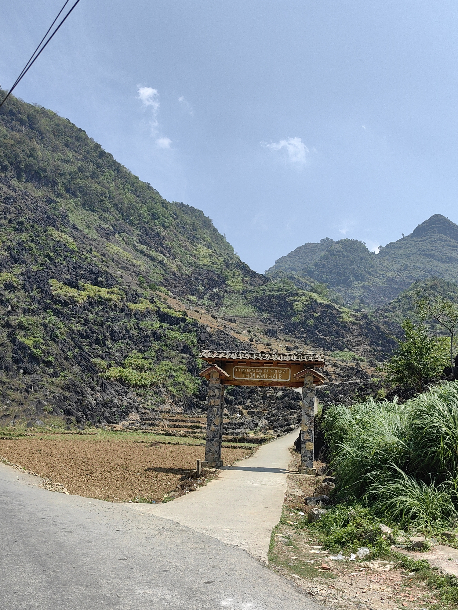The image shows a rural scene in Yên Minh Commune, Vietnam. In the foreground, there's a paved road intersecting with a narrower concrete path that leads through a stone and tiled gate. The gate bears signage in Vietnamese. To the side of the path, there is a cultivated field of brown soil. The right side of the path has dense green vegetation. The background reveals a rugged mountain landscape covered in sparse green vegetation and exposed black rock formations. The sky is clear blue with a few scattered clouds. An overhead power line is visible in the upper left corner.