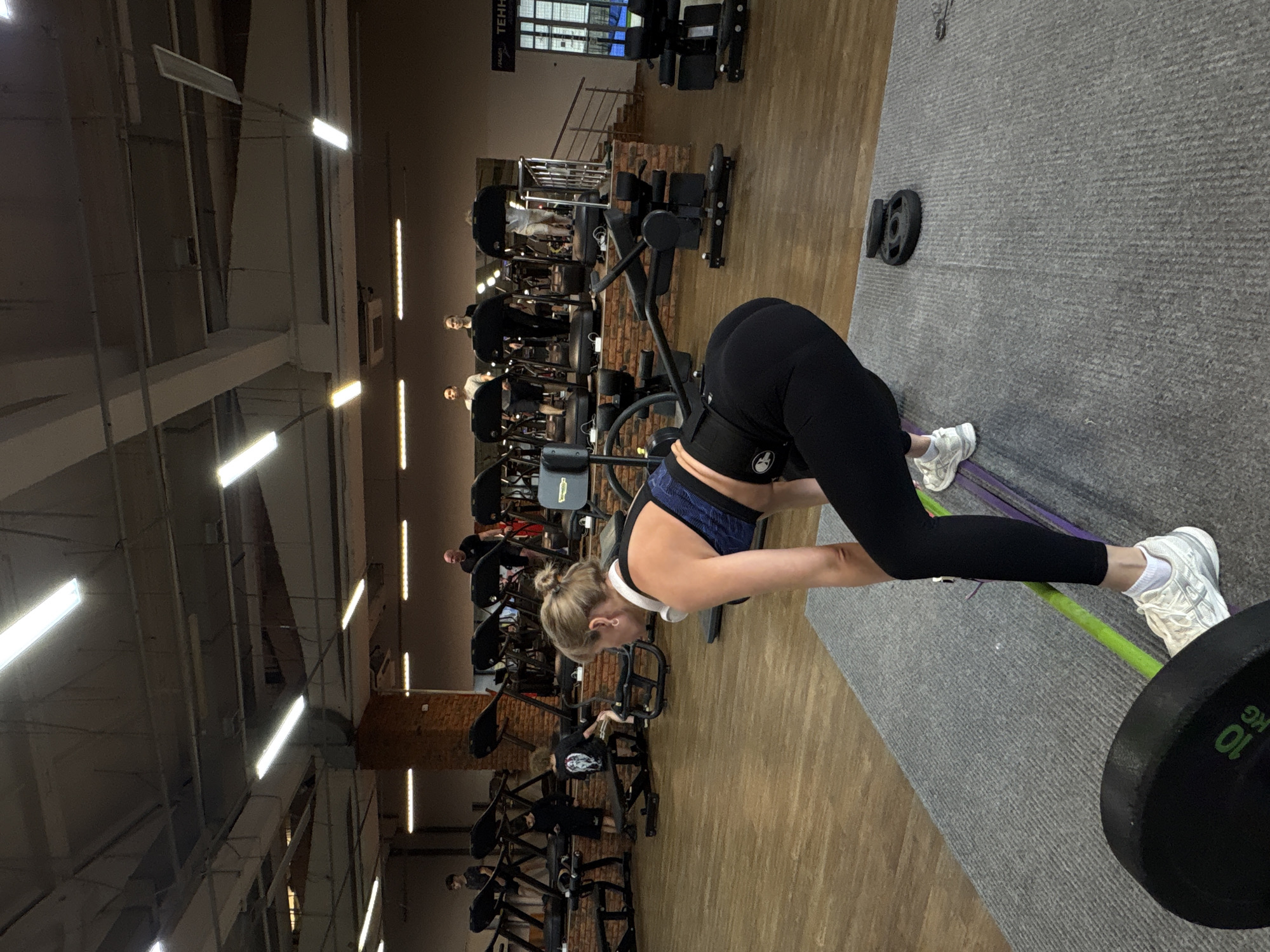 In a fitness center in Chaikovsky, Russia, a woman is exercising with a barbell and resistance band. She is wearing a sports bra, black leggings, and white sneakers. The barbell has a 10 kg weight on one side. Other gym equipment and people can be seen in the background. The floor is a combination of wood and gray carpet. Artificial lights illuminate the scene.