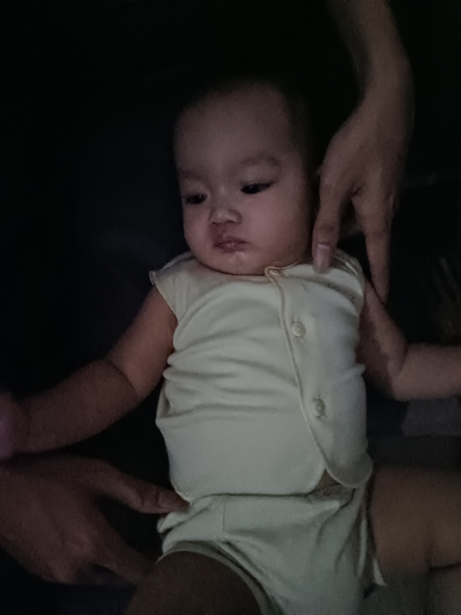 The image shows a close-up of a baby wearing a light-colored sleeveless onesie and shorts. The baby has dark hair and is looking towards the left of the frame with a slightly pouting expression. Two adult hands are visible in the frame, one near the baby's head and another near the baby's legs, suggesting the baby is being held or supported. The background is dark and appears to be indoors. The image does not contain any elements that indicate a specific location in West Jakarta, Indonesia.