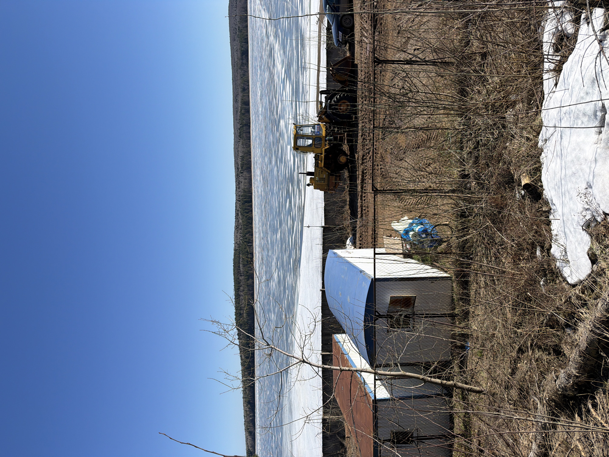 The image depicts a partly frozen body of water, likely a reservoir, bordering a wooded area and a developed area with buildings and equipment. A yellow tractor is parked near the water's edge, and a car is nearby. A small, white building with a curved roof is visible. The environment appears to be in a state of transition from winter to spring, with patches of snow and bare trees. The clear blue sky suggests a sunny day. Without additional information, it is difficult to confirm the exact location in Chaikovsky, Russia, but the scene is likely near the Kama reservoir.