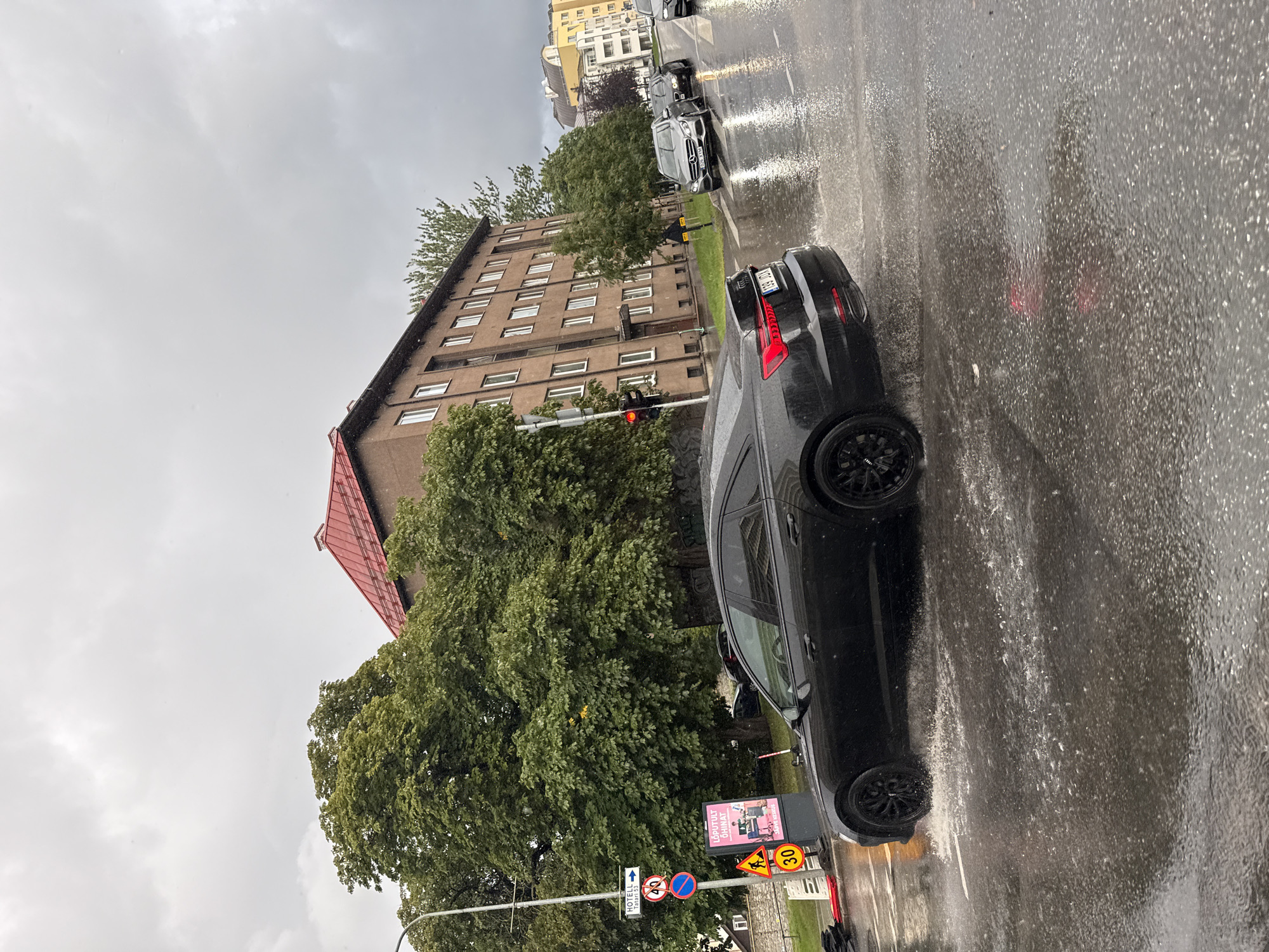 The image depicts a street scene in Tallinn, Estonia, during what appears to be heavy rain. A black Audi sedan is driving through a large puddle on the wet asphalt road, creating significant spray. In the background, there is a multi-story brown building with a red roof and numerous windows. Trees with lush green foliage are visible on the left side of the building. A traffic light with a red signal is mounted on a pole on the left side of the street. Various traffic signs are also present, including one indicating "HOTEL" and a speed limit sign showing "30". Another sign displays "LÕPULT ÕHNAT". Several other vehicles, including a silver Mercedes-Benz SUV, are present further down the street. The sky is overcast and gray, consistent with rainy weather. The overall environment suggests an urban setting experiencing inclement weather conditions.