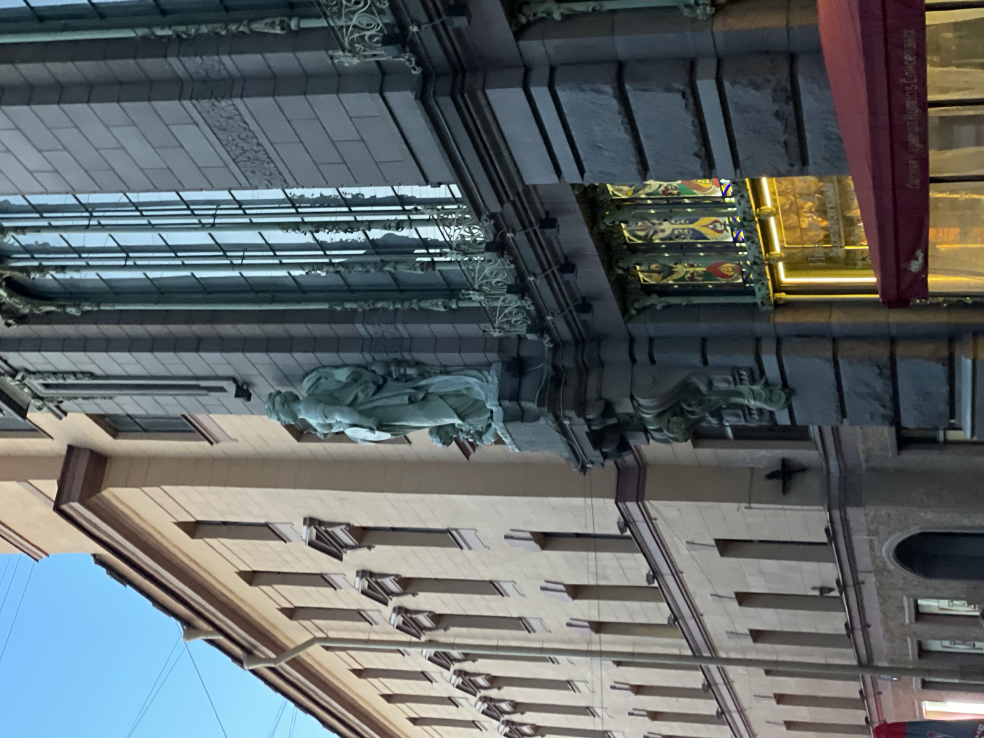 This daytime image, captured under a clear blue sky, presents an upward view of two adjacent building facades in an urban setting. The main subject is an ornate, multi-story building, likely a historic department store, characterized by its elaborate architectural details.
The upper part of this building features a distinctive glass and dark metal canopy or roof structure, possibly covering an atrium, adorned with intricate wrought-iron decorative elements. Below this, the facade is constructed from dark grey stone or brick. A prominent classical statue of a female figure is affixed to this dark wall.
At street level, an illuminated shopfront is visible, embellished with colorful decorative panels or stained glass above the entrance. A red awning or sign hangs vertically, displaying Russian text: "Русская кухня и Деликатесы" (Russian cuisine and Delicacies) and "Gastronom N°1". This identifies the establishment as a specialty food store. Faintly visible text inside the shop may include "УНИВЕРМАГ" (Department Store).
To the right of this central edifice, a portion of a lighter-colored, less ornate building is visible. Its facade, possibly made of beige stone or stucco, features rows of rectangular windows with pronounced sills or lintels creating a shadow pattern.
The architectural style, ornate detailing, and the visible Russian text strongly suggest the location is the historic Eliseev Emporium (Eliseevsky Gastronom) in Saint Petersburg, Russia, offering a glimpse into the city's grand architectural heritage and commercial life.