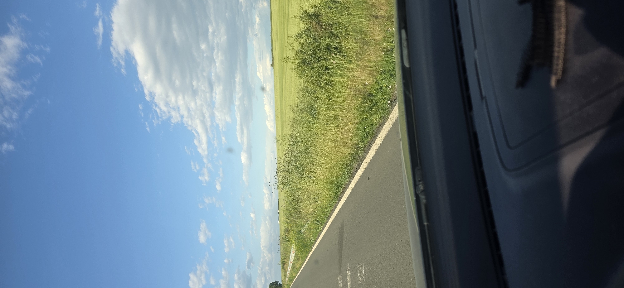 The image depicts a view from inside a vehicle, likely a car, moving along a rural road. Outside the window is a clear, blue sky with scattered cumulus clouds. A field of green grass and vegetation borders the road. A black-colored car doorframe is visible on the right edge of the image, creating a framed view of the landscape. The location is Troyanovo, Bulgaria based on available GPS metadata.