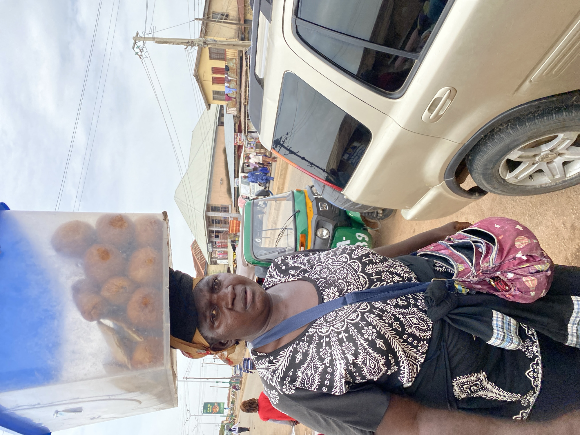 An elderly woman stands on a street in Bukuru, Nigeria, carrying a plastic container filled with round pastries on her head. She wears a black and white patterned top and has a shoulder bag. A gold-colored SUV is parked behind her, and a green and yellow three-wheeled vehicle is visible further down the street. Buildings line the street, with clothes hanging on a line between them. A utility pole stands in the background, and other pedestrians are visible in the distance.