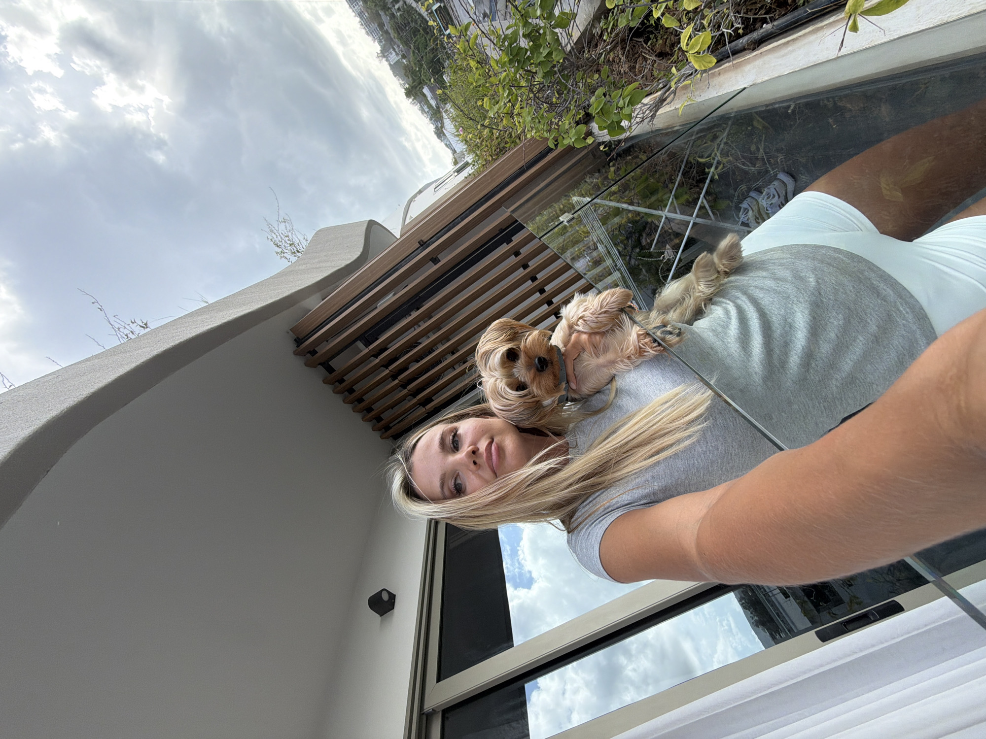 A blonde woman with a Yorkshire terrier is standing on a balcony in Choeng Thale, Thailand. The woman is wearing a grey t-shirt and white shorts, and she is holding the dog in her arms. The dog is looking at the camera with its tongue sticking out. The balcony has a glass railing and a wooden slatted screen. In the background, there is a view of the city with buildings and trees. The sky is cloudy.