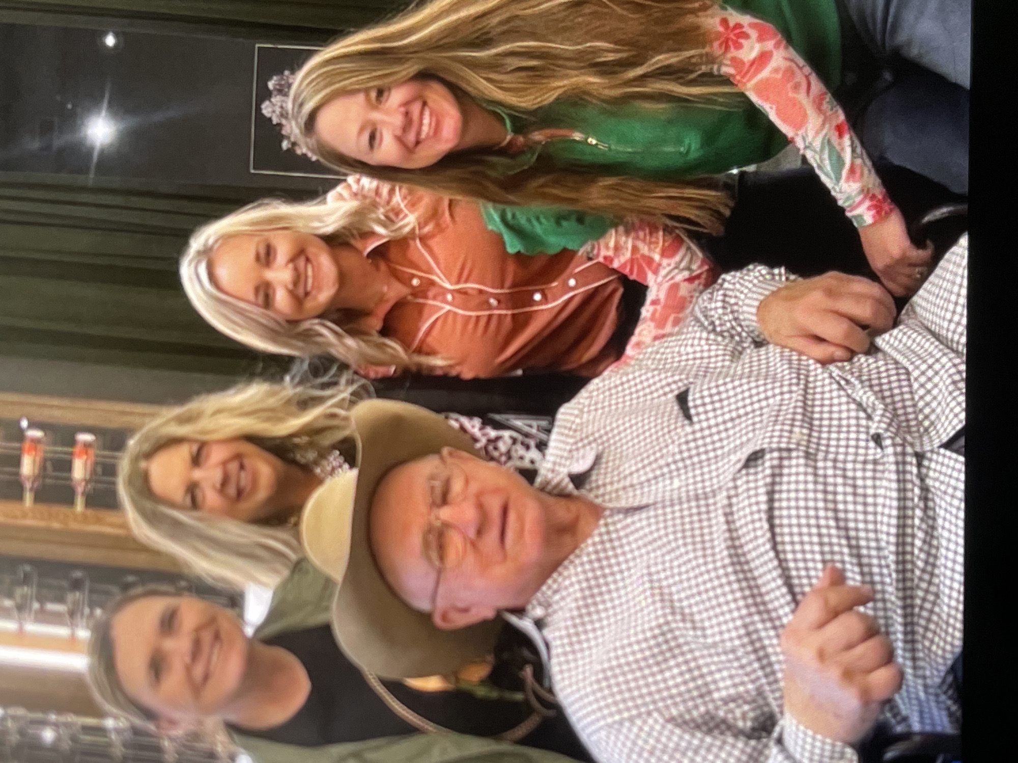 A medium shot captures a group of five people, four women and one man, posing for a photograph indoors. The man in the foreground, seated and wearing a cowboy hat and a checkered shirt, is positioned to the left. The four women are standing behind him. The woman on the far left has short hair and is wearing a green jacket over a black shirt. Next to her is a woman with long blonde hair and a patterned necklace. In the center, a woman with blonde hair is wearing a rust-colored shirt with white piping. On the far right, a woman with long blonde hair wears a green top with ruffled sleeves and a floral-patterned shirt underneath. The background is slightly out of focus and appears to be an interior space with wooden shelves and dark green curtains. There are no discernible geographical markers to indicate a specific location within the city of Unknown, Unknown.