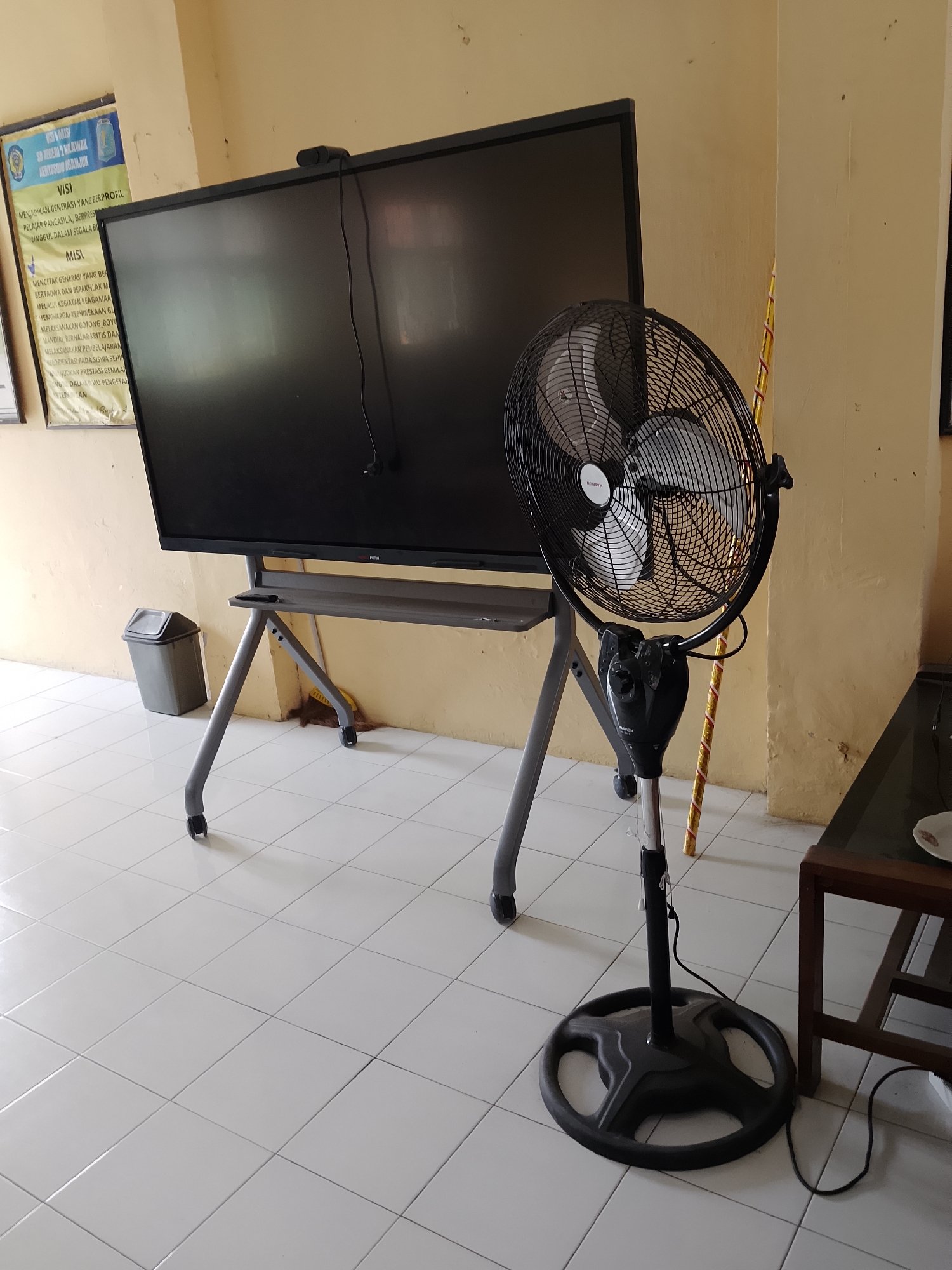 The image shows an indoor scene, likely a classroom or office space, with a large, black, modern display screen mounted on a movable gray stand. To the right of the screen, a black oscillating floor fan stands on a circular base, with its power cord visible on the floor. A small gray trash can is situated to the left of the display screen stand. The floor is tiled with white square tiles. In the background, a yellow wall is visible, with a poster hanging on it. The poster, partially visible on the left, appears to be informational, with text in Indonesian, including the words "VISI" and "MISI". The presence of a display screen, a fan, and educational posters suggests an institutional or educational setting. The environment is clean and well-lit, with natural light coming from an unseen source. There are no people visible in the image.