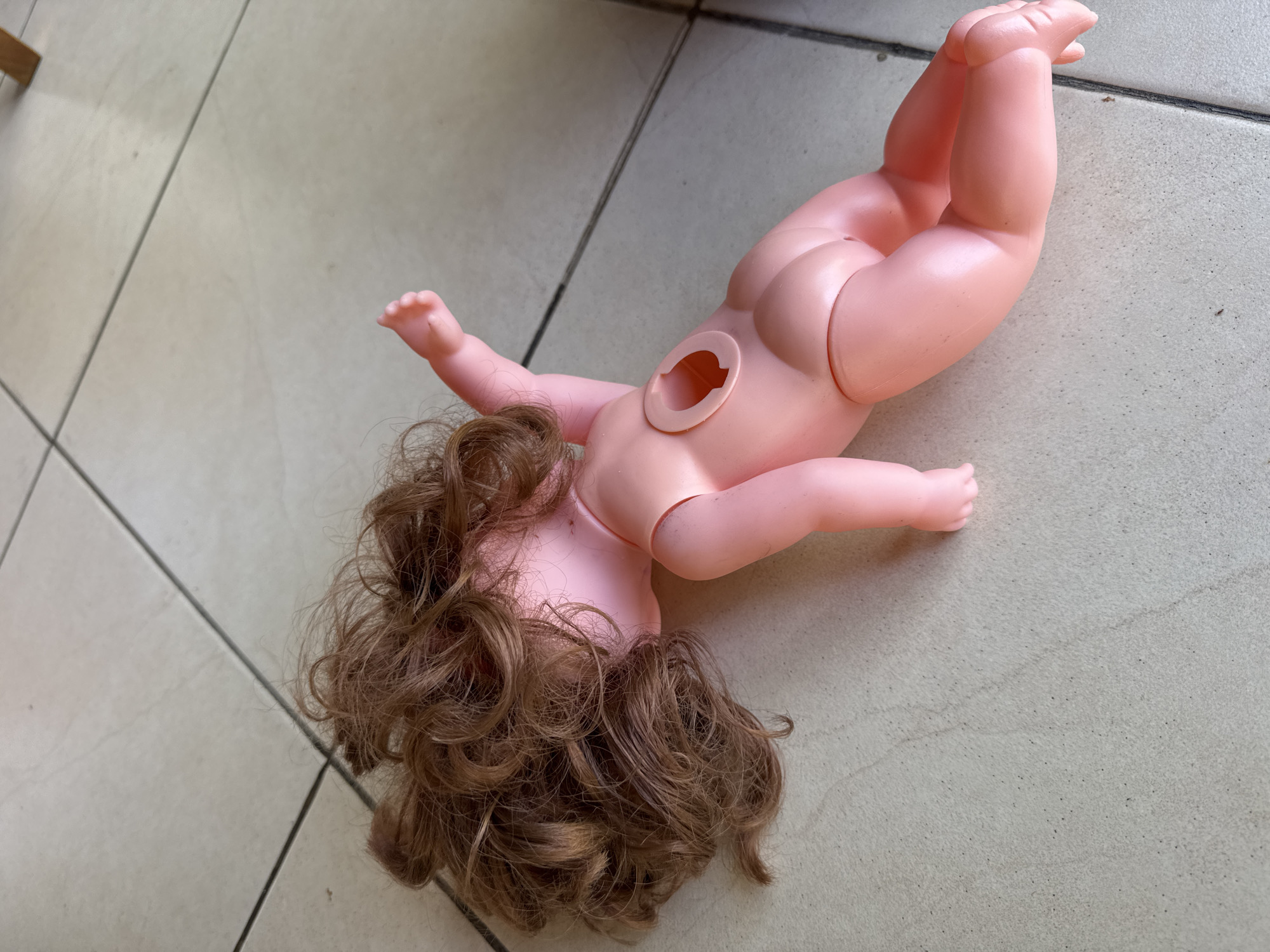 A nude, pink plastic doll lies on a tiled floor. The doll is missing its head, which has been detached and is oriented so that its brown, curly hair is facing downward. The doll's body has a circular opening in the center of its back where the head was attached. The floor tiles are light-colored and appear to be located indoors. The photograph was taken in Kuta Selatan, Indonesia.