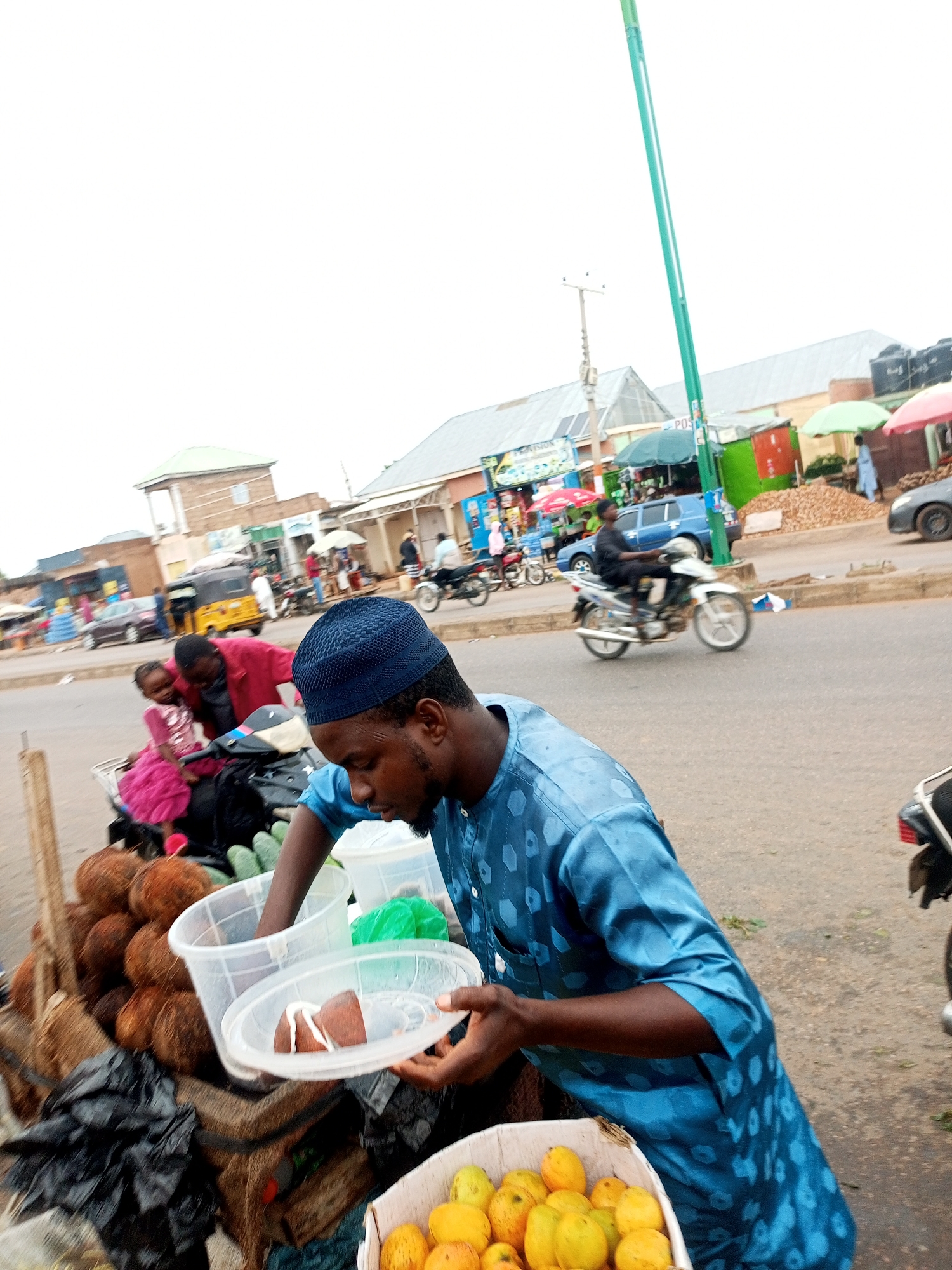 A man in a blue patterned garment and kufi cap is preparing food items in a market stall in Tempure, Nigeria. He is holding a clear plastic container with two brown food items topped with white cream. In the foreground, a large basket of yellow fruits and coconuts are visible. Behind him, a young girl in a pink dress is seated on a motorbike. The market scene extends to the street where other people, vehicles including a blue car and motorcycles, and various market stalls with umbrellas are present. The sky is overcast.