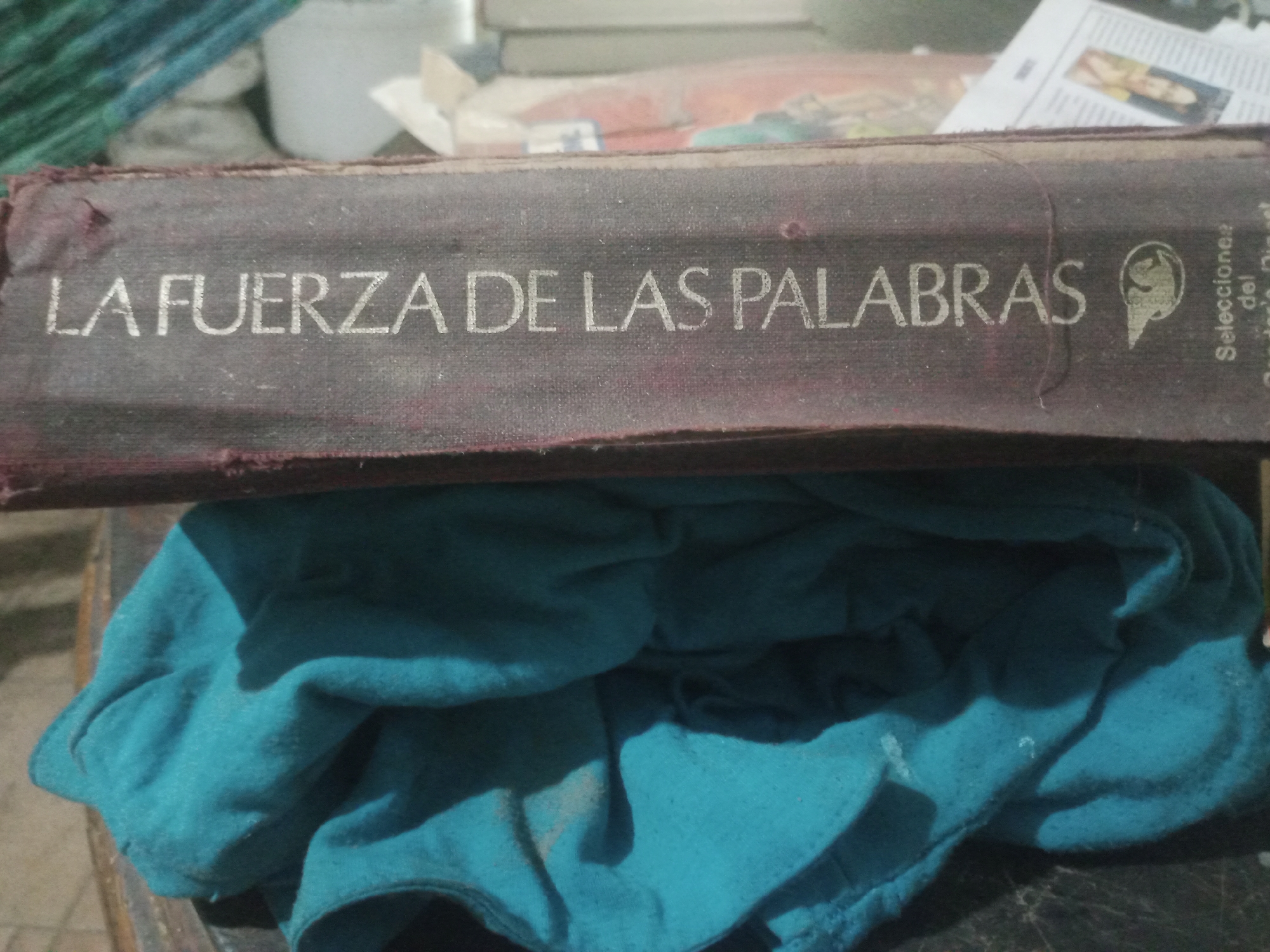 This is a close-up, medium-angle shot of a book spine that reads "LA FUERZA DE LAS PALABRAS" in a light, sans-serif font. The book's cover is a dark, textured material, possibly fabric or aged leather, with visible dust. To the right of the title, there is a circular emblem containing a silhouette of a bird within a crest. Further to the right, text is visible vertically: "Selecciones" above "del". The book is resting horizontally on a surface, and below it, a crumpled teal-colored cloth occupies the lower half of the frame, casting shadows. The background is blurred but suggests a cluttered environment with other books and objects, possibly in a home setting. The scene is located in the city of Santa Ana, Venezuela.
