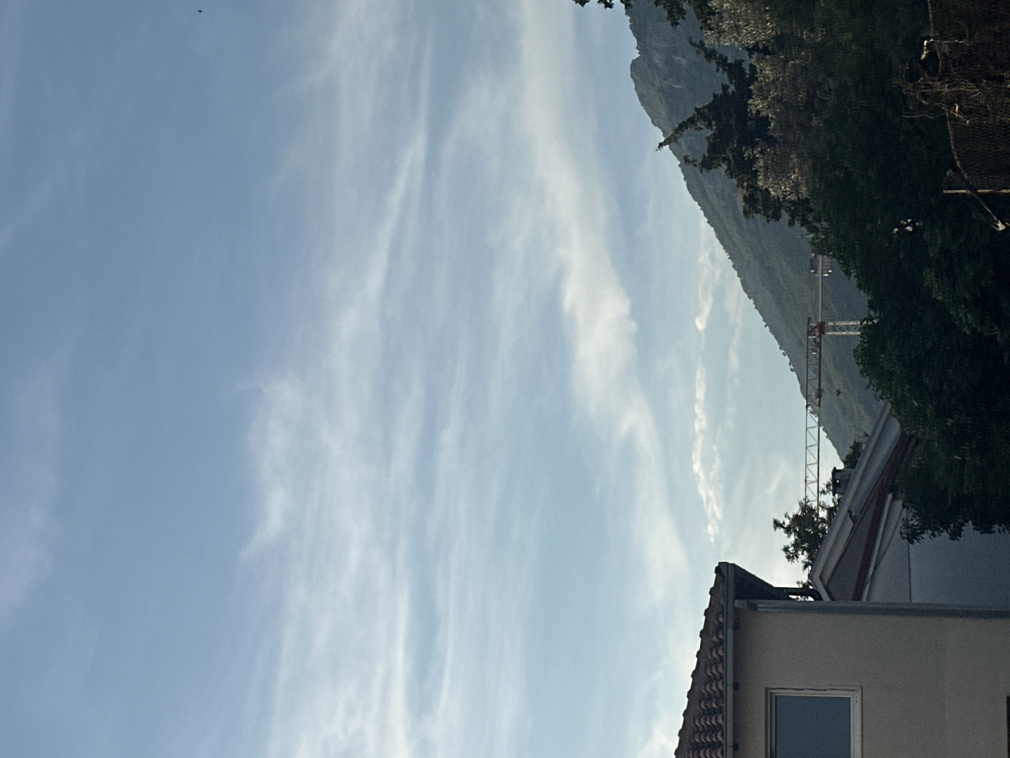 The image depicts a daytime scene in Bar, Montenegro, with a light blue sky filled with elongated cirrus clouds. A mountain covered in vegetation rises on the right side of the frame, behind a partially visible construction crane. In the lower right corner, a building with a tiled roof and a window is also visible. The environment appears urban, bordering a mountainous area. No people are visible.