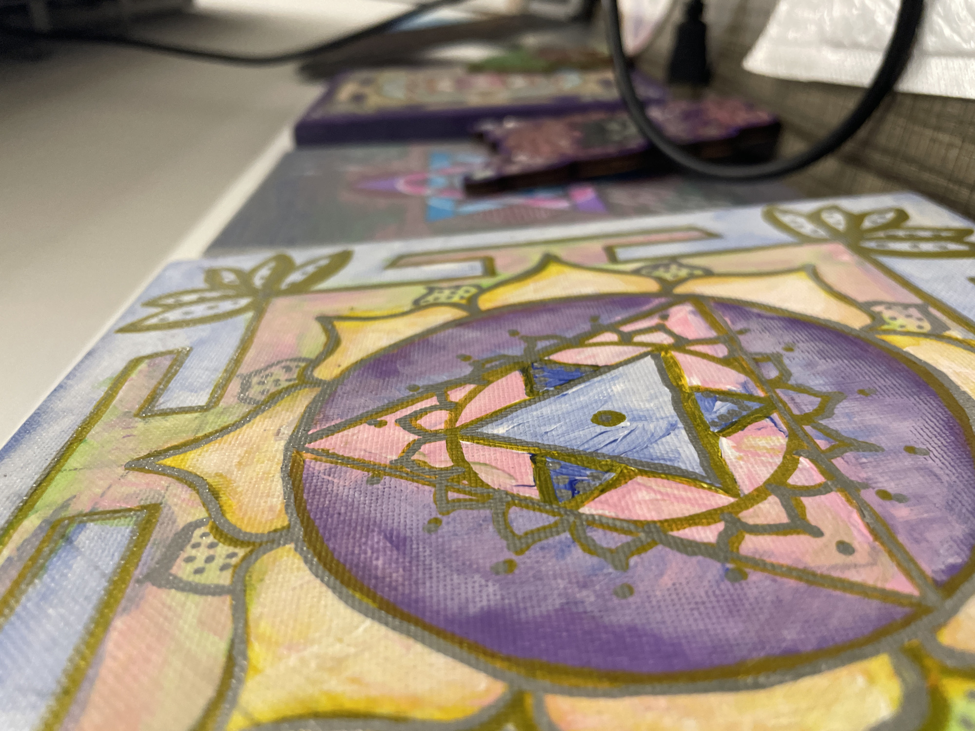 The image displays a close-up, shallow depth-of-field view of a vibrant, hand-painted artwork, likely on a small canvas. The central subject, in sharp focus, is a square painting featuring a geometric, mandala-like design. It has a prominent purple circular core containing intricate blue and pink triangular shapes, all outlined with a shimmering metallic gold or silver paint. Surrounding the circle are sections of pastel yellow and light blue/purple, also outlined in metallic paint, depicting stylized floral or leaf patterns. The texture of the canvas and visible brushstrokes are clearly discernible.
In the blurred background, several other similar artworks are arranged in a receding line, suggesting a series or collection of these pieces. A black cable or wire is visible in the upper right quadrant and extending into the background, alongside a blurry white object. The setting appears to be indoors, with the artworks resting on a light-colored surface, possibly a table or shelf. The lighting is bright and even, illuminating the painting's colors and details. No specific activity is taking place, but the image highlights completed or ongoing creative work. No text is visible in the image. The location context provided is Bac Nha Trang Ward, Vietnam.