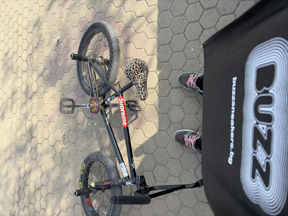 The image captures a top-down view of a black BMX bicycle lying on light-colored hexagonal paving stones, observed from the perspective of an individual standing over it. The scene is illuminated by direct sunlight, casting distinct shadows. The location is an outdoor paved area in Sofia, Bulgaria, indicated by the "buzzsneakers.bg" text on a visible bag.
In the foreground, a black bag or object is partially visible, displaying a stylized white "BUZZ" logo and "buzzsneakers.bg" written in white text, oriented upside down relative to the viewer. Immediately behind this, the feet of an individual are visible, wearing dark-colored sneakers with light pink laces and a white sole. The shoes feature a light blue or teal accent, possibly a brand swoosh.
The central subject is a black BMX bicycle. Its frame has a prominent red "Supreme" sticker on the downtube and several other stickers, including one that appears to say "THE" near the head tube. The bicycle is equipped with a leopard-print seat and a multi-colored, iridescent sprocket. Black pedals and handlebars with black grips are visible. The wheels are black, and one tire displays yellow branding. A black stand or support arm is attached near the front wheel.
The ground consists of hexagonal paver blocks in varying shades of light gray and beige. A small piece of red and white litter is present on the paving in the upper right portion of the frame.