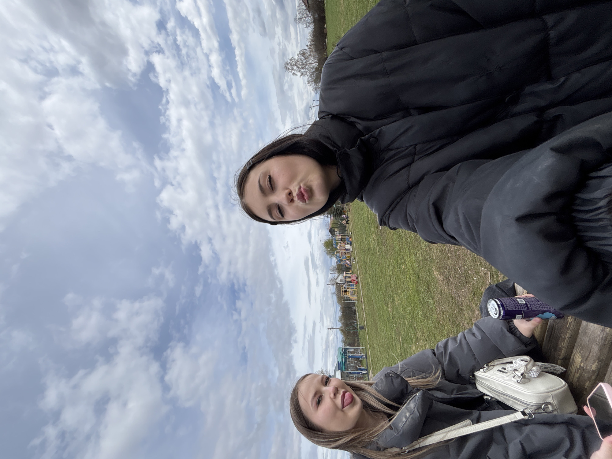 Two women are posing for a picture outdoors, in Фоки, Russia, with a cloudy sky overhead. The woman in the foreground sticks out her tongue, wearing a grey puffer jacket. A white purse, a phone, and a purple drink can sit next to her. The woman in the background wears a black puffer jacket and puckers her lips. In the background, there is a grassy area with a playground.