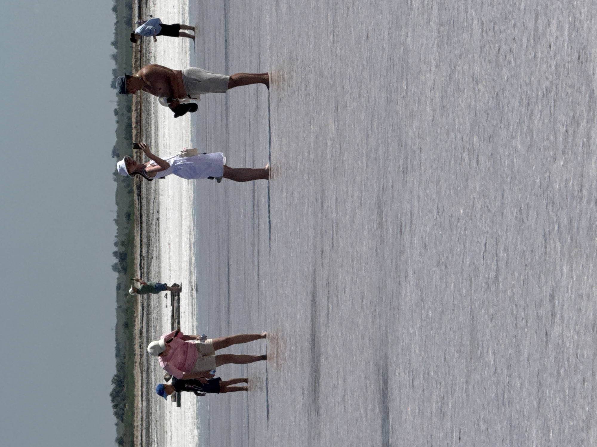 This image depicts a group of people standing in shallow, reflective water, possibly a salt lake or salt flats, with a treeline in the background. The sky is overcast and pale. In the foreground, a man in a dark hat and light-colored shorts stands looking towards a woman in a white dress and hat who is taking a photo with her phone. To their left, a young boy in a blue cap and dark shirt walks ahead of a woman in a pink shirt and light-colored shorts, both holding plastic bottles. Further in the background, another person in a light blue shirt stands in the water, and a person in a green shirt and shorts is visible on a wooden structure. The environment appears to be a bright, open expanse of water with a rough texture. The location is identified as Bursol', Russia.