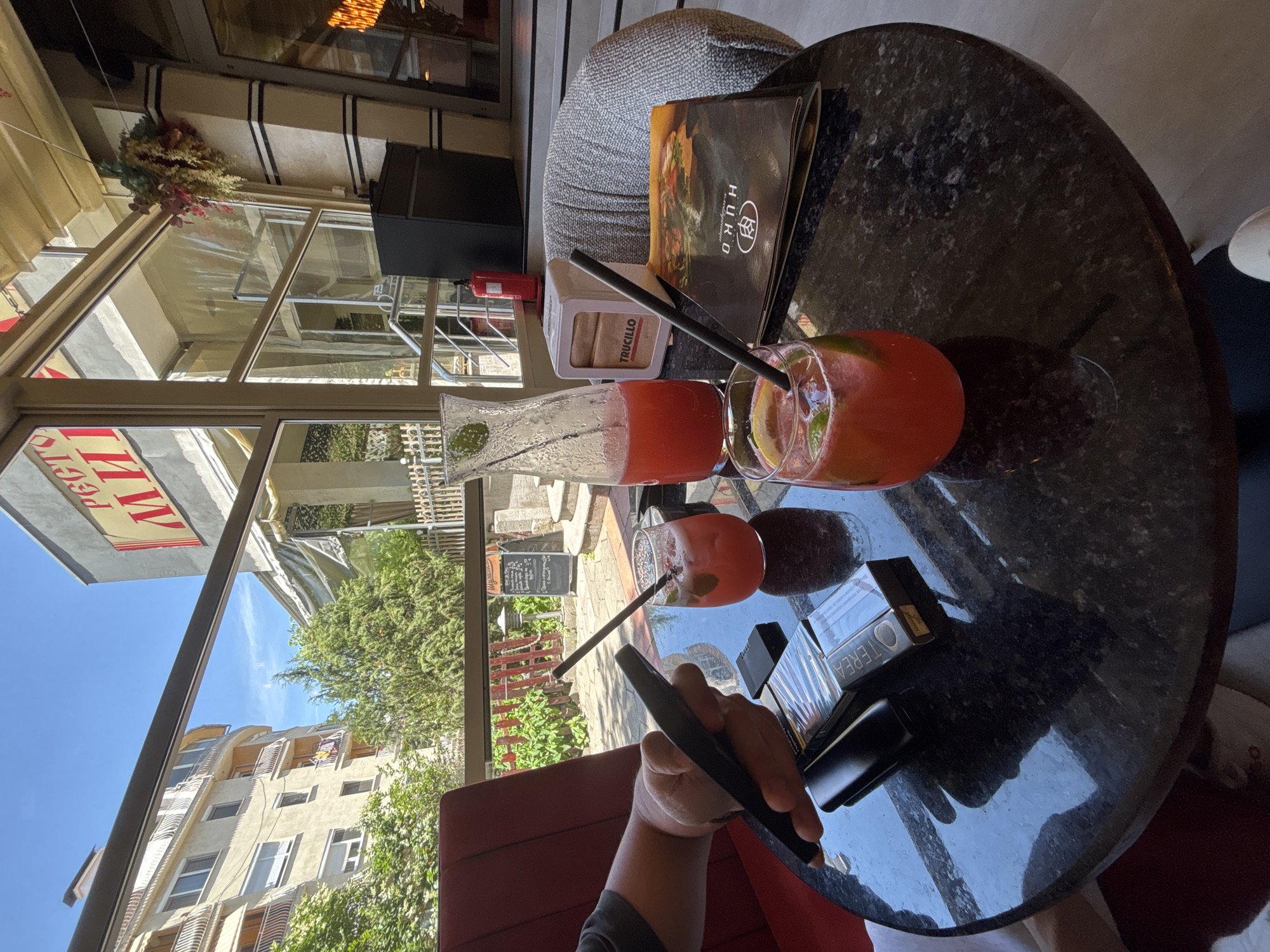 A person's hand is holding a black mobile phone over a dark, speckled table. To the left of the phone is a pack of cigarettes and a black electronic cigarette device. In front of the phone are two glasses filled with a red beverage, garnished with lime and mint, and each with a black straw. A glass carafe containing the same red beverage is to the right of the glasses. A white napkin dispenser with the word "TRUCILLO" printed on it sits between the carafe and a grey upholstered chair. To the right of the chair is a folded menu with an image of food. The scene is set outdoors at a cafe, with a view of a building and trees through a glass window. In the background, above the window, a sign with red lettering is visible, reading "РЕСТО МИ." This cafe is located in Plovdiv, Bulgaria.