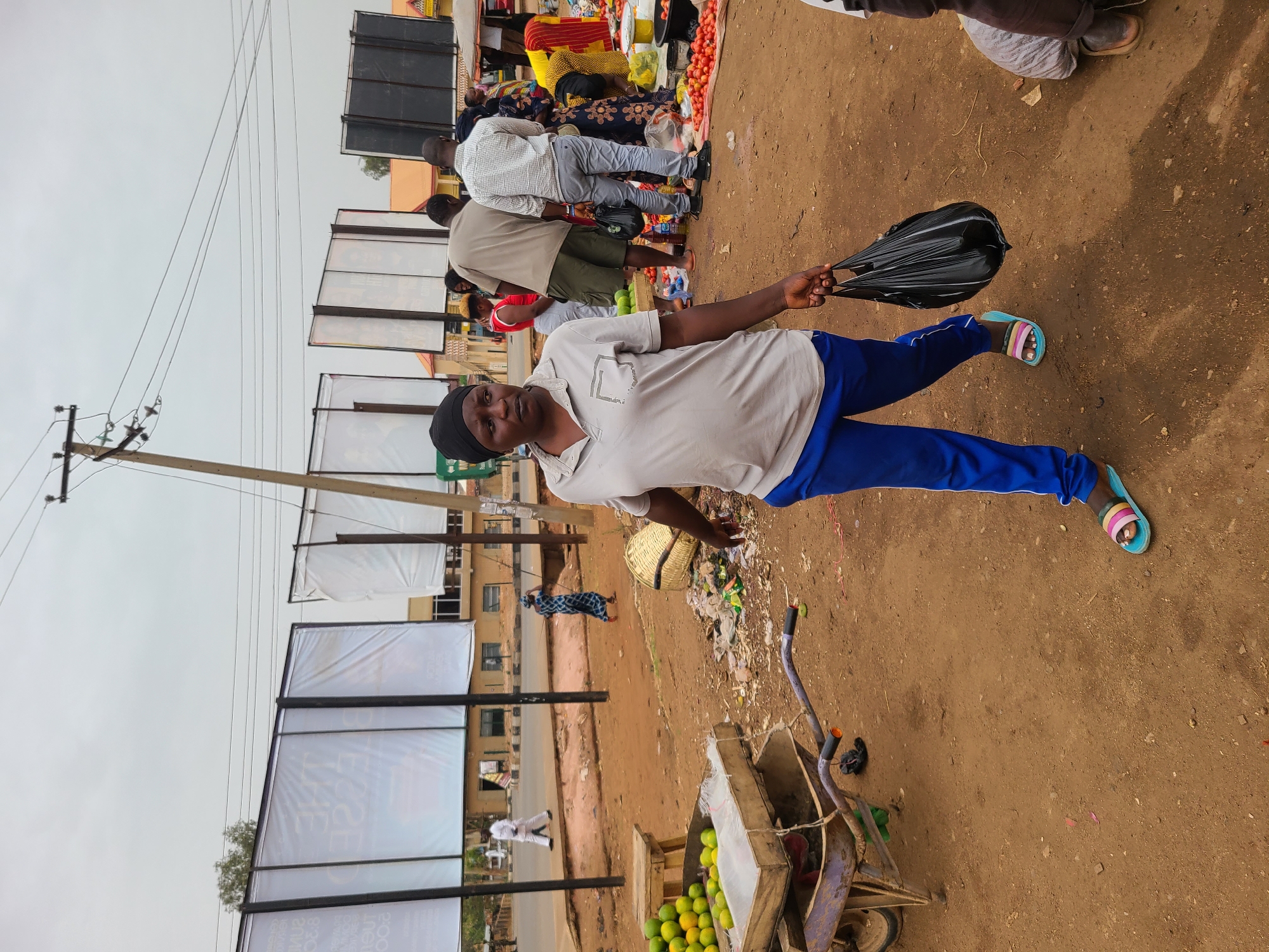 In a medium-angle shot taken in Jos, Nigeria, a man wearing a light gray polo shirt, blue track pants, a black head covering, and colorful sandals, stands on a reddish-brown dirt patch. He holds a black plastic bag in his right hand. Behind him, people are gathered at what appears to be a market stall selling tomatoes and other goods. To the left of the man are a series of advertisement billboards, some with white material covering the displayed ads, against a cloudy sky. A utility pole is also visible. In the foreground, there is a wheelbarrow with a wooden crate filled with green fruit. Garbage is strewn near the wheelbarrow.