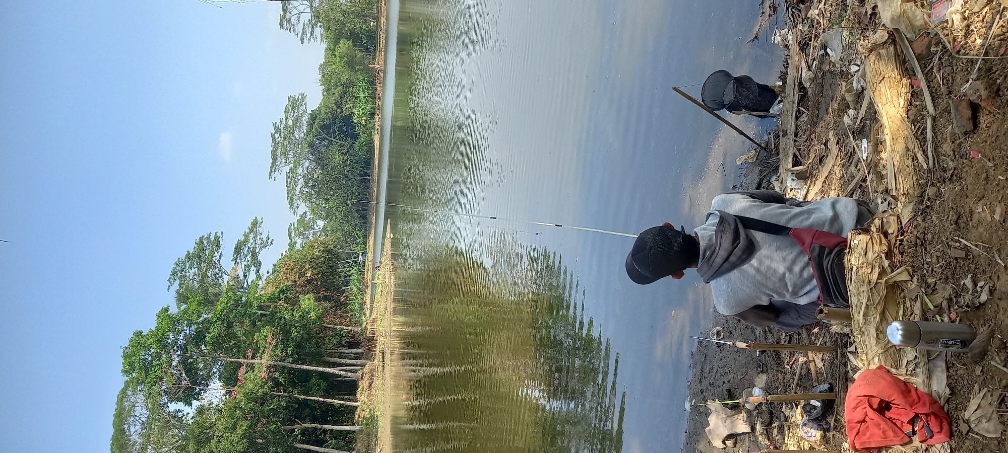 In Sidamulya, Indonesia, a person wearing a grey hoodie and a black cap sits on the muddy bank of a calm lake, facing away from the camera. They are fishing, with a fishing rod extended over the water. Several other fishing rods are planted in the ground to their left. To the right of the fisherman, a fishing net is propped up by a stick. The surrounding environment is natural, with lush green trees lining the opposite bank of the lake. Debris, including plastic bags and pieces of wood, are scattered on the ground near the fisherman. The sky is clear and blue.