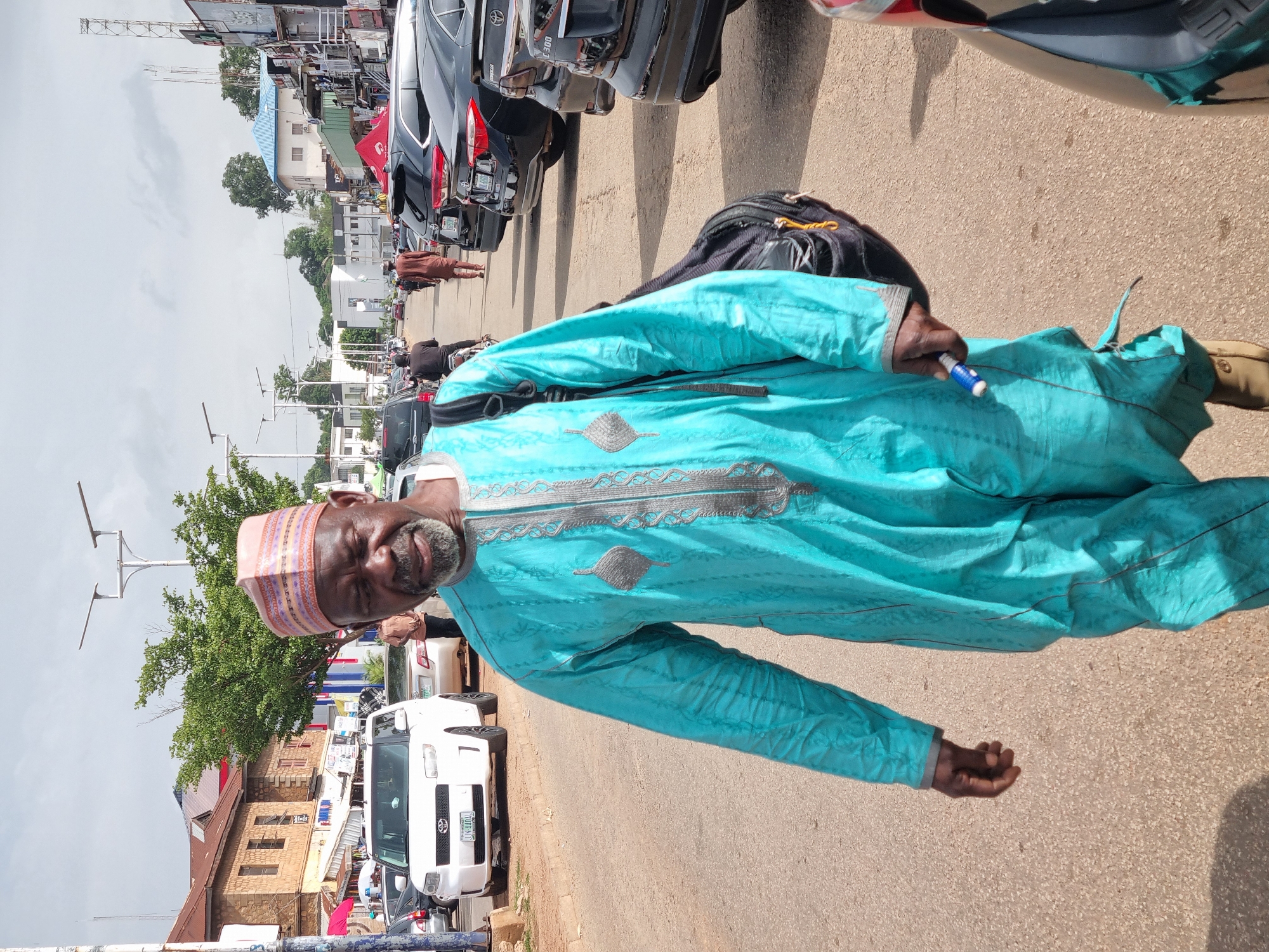 A man in a teal traditional garment stands on a paved street in Jos, Nigeria, facing the camera and looking to his left. He has a gray beard and is wearing a patterned cap. He is holding a white object with a blue tip in his right hand. Behind him, a black backpack rests on the ground. The street is lined with parked cars, including a white Toyota vehicle with the license plate "JN-840A4". In the background, buildings and trees are visible. The sky is overcast, suggesting a cloudy day. The image appears to have been taken during daylight hours.
