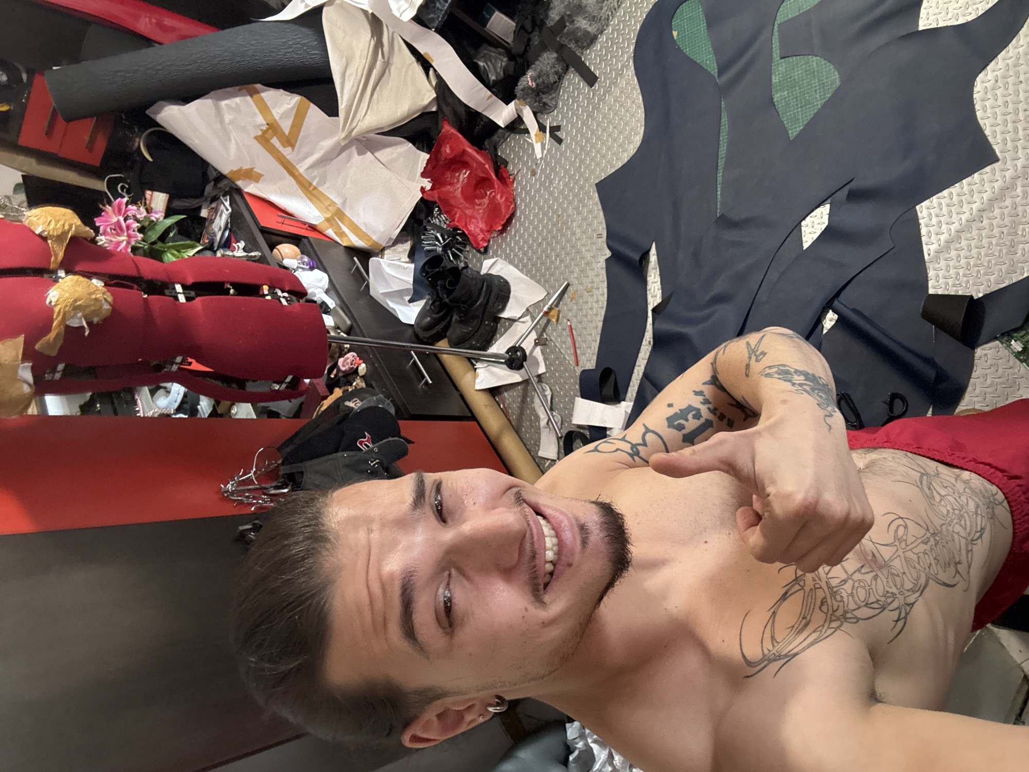 A shirtless man with dark hair pulled back, a goatee, and extensive tattoos on his chest and arms, is smiling and giving a thumbs-up gesture in what appears to be a workshop or studio space. He is wearing red shorts. In the background, there are various items related to crafting or sewing, including a red mannequin, rolls of fabric, and cut pieces of dark material laid out on a textured metal floor. The environment suggests a creative workspace.