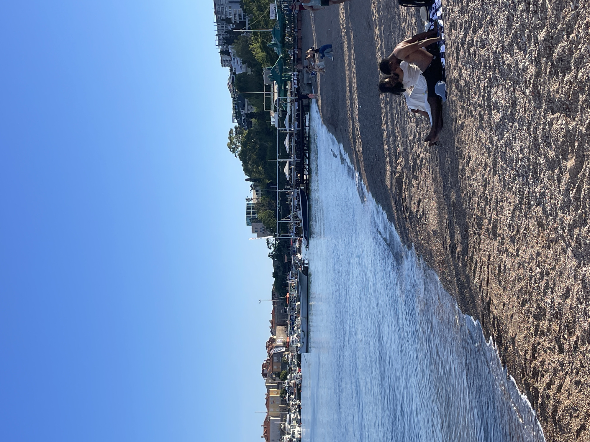 This image captures a coastal scene, likely in Budva, Montenegro, given the architecture and setting. On the right, a sandy beach stretches along the water's edge, where a couple stands. The individual on the left is clad in a white garment, while the individual on the right is shirtless and wearing dark shorts. The duo appears to be interacting intimately, possibly embracing or kissing. A towel lies beneath them on the sand.
To the left of the beach is a body of water, filled with boats and docks. Beyond the water, buildings and vegetation line the shore, forming a cityscape. The sky above is clear and blue. There are some other people walking on the beach but they are further in the distance.