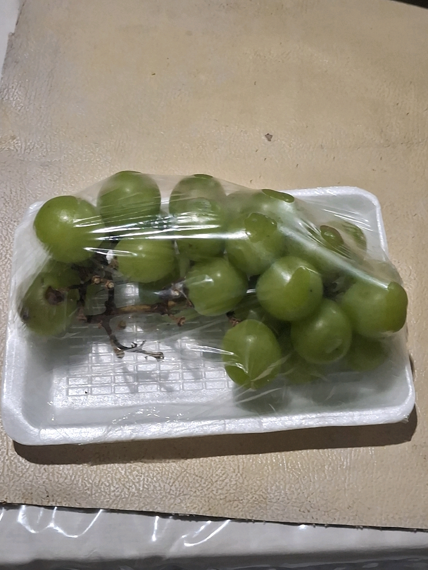 A bunch of green grapes is packaged in plastic wrap and presented on a white styrofoam tray. The grapes are clustered together, with some showing slight imperfections. The tray is placed on a textured surface, likely a countertop or table. The lighting suggests an indoor setting, and the overall scene is straightforward, focusing on the fruit packaging. The image does not provide any information about its location in Lumajang, Indonesia.