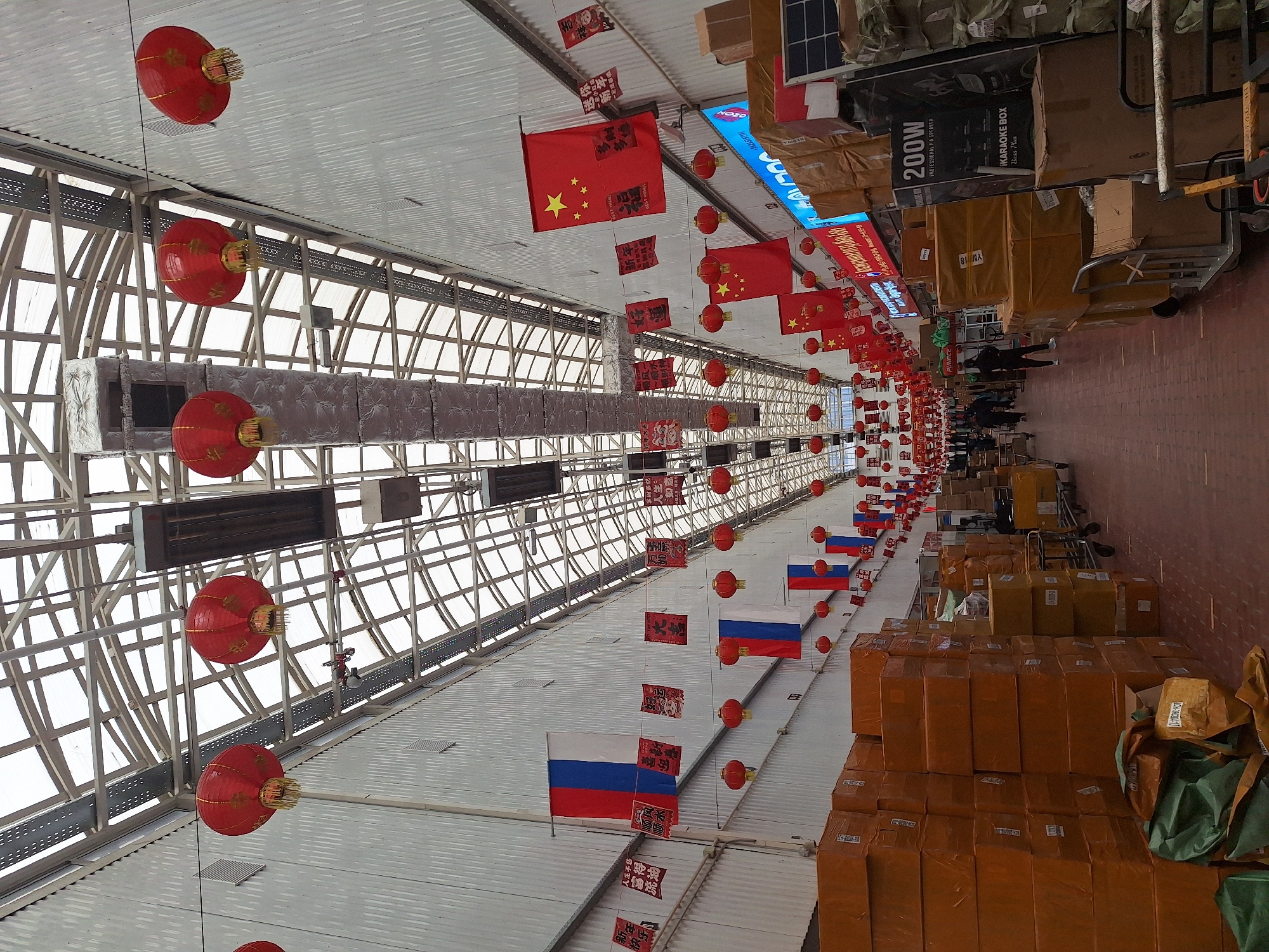Here is a detailed description of the image:
**Scene:** The image captures an indoor market or warehouse in what seems to be Moscow, Russia, due to the Russian flags displayed alongside Chinese flags. The location is brightly lit by natural light filtering through a lattice-like window structure on the left. The scene is decorated with red Chinese lanterns and banners featuring Chinese characters, suggesting a festive or celebratory event, potentially Chinese New Year.
**Objects:** The interior is filled with numerous cardboard boxes, likely containing merchandise, stacked high on the right side of the frame. There is a dolly/cart loaded with boxes. Additionally, there are packages wrapped in brown paper and some green bags.
**People:** A group of people are walking through the warehouse space.
**Environment:** The overall environment is busy, cluttered, and functional. The focus is clearly on commerce and storage.